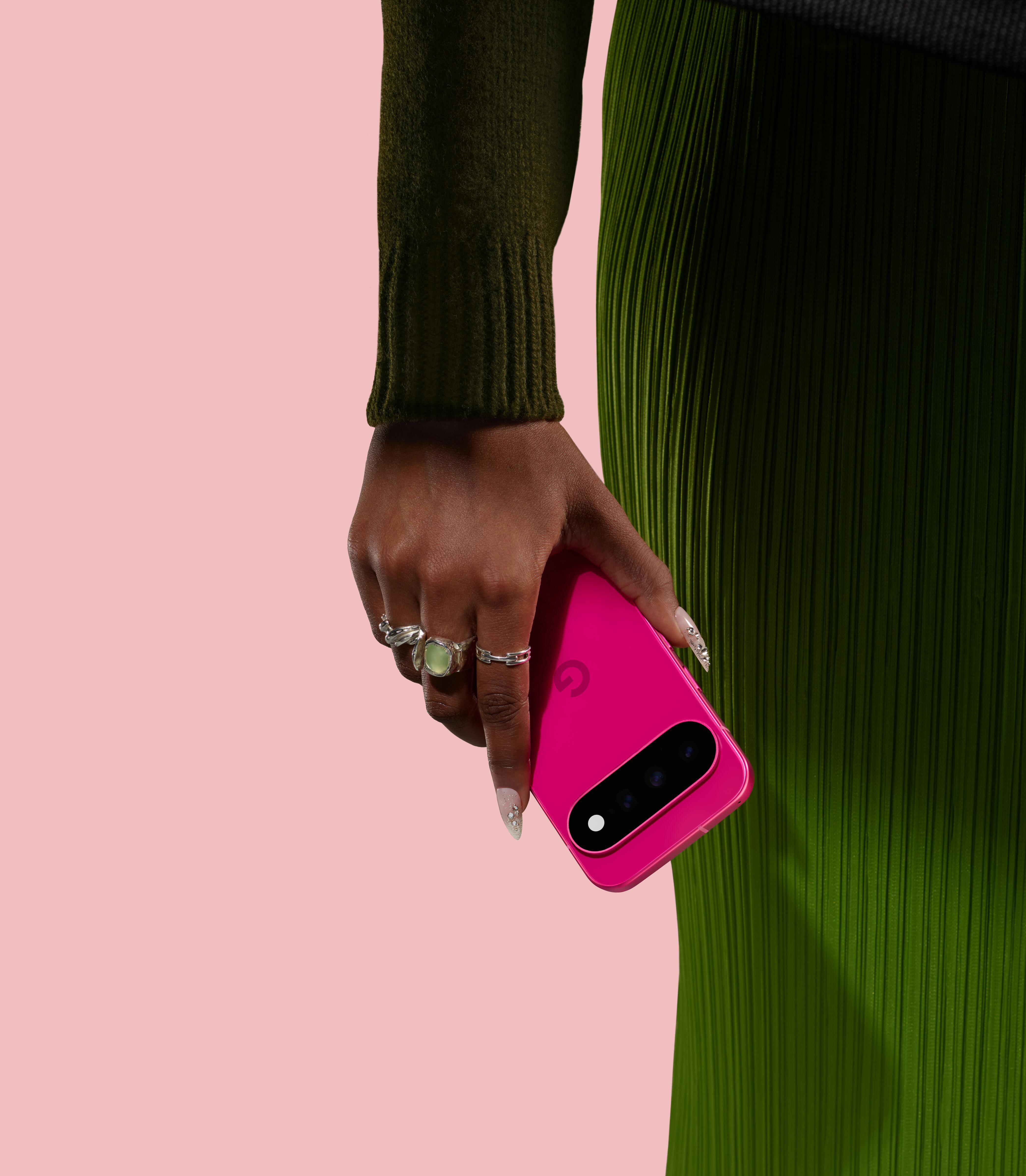 Eine Person in grüner Kleidung und mit Ringen hält ein hellrosa Smartphone vor einen rosa Hintergrund.