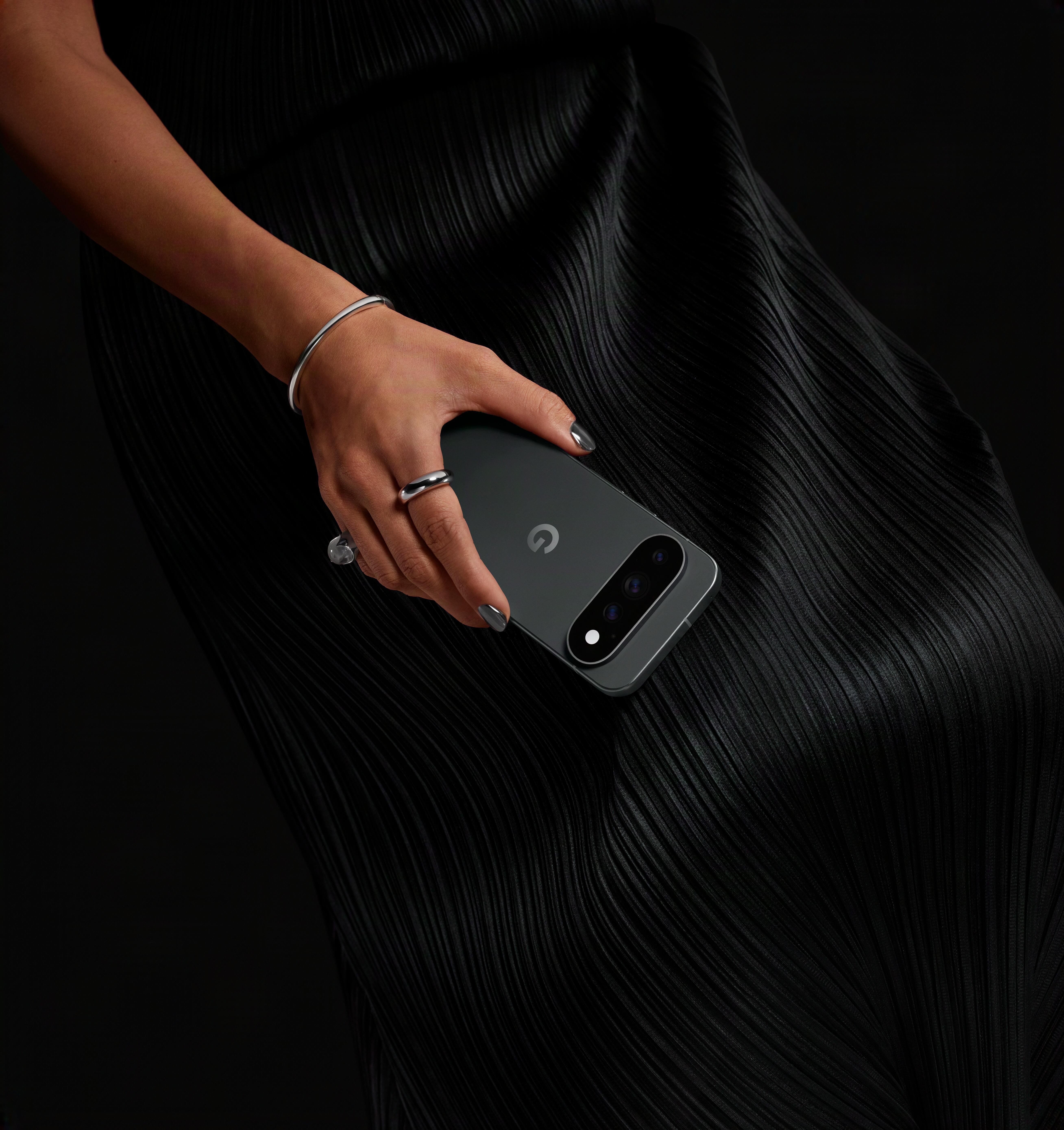 Eine Person in einem schwarzen Plissee-Kleid hält ein dunkelgraues Smartphone mit Google-Logo vor dunklem Hintergrund.