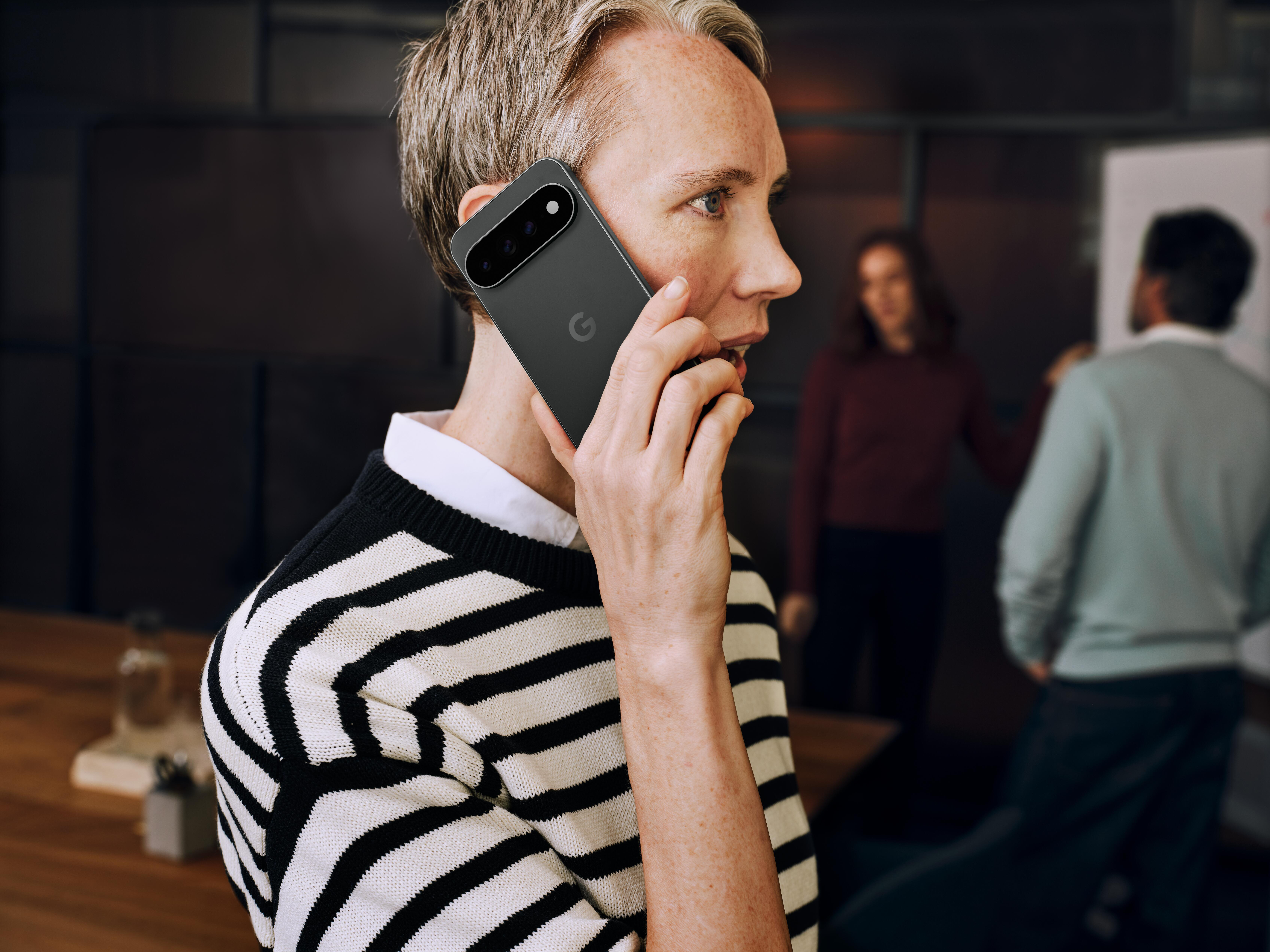 Eine Person im gestreiften Pullover telefoniert im Büro, im Hintergrund zwei verschwommene Personen.