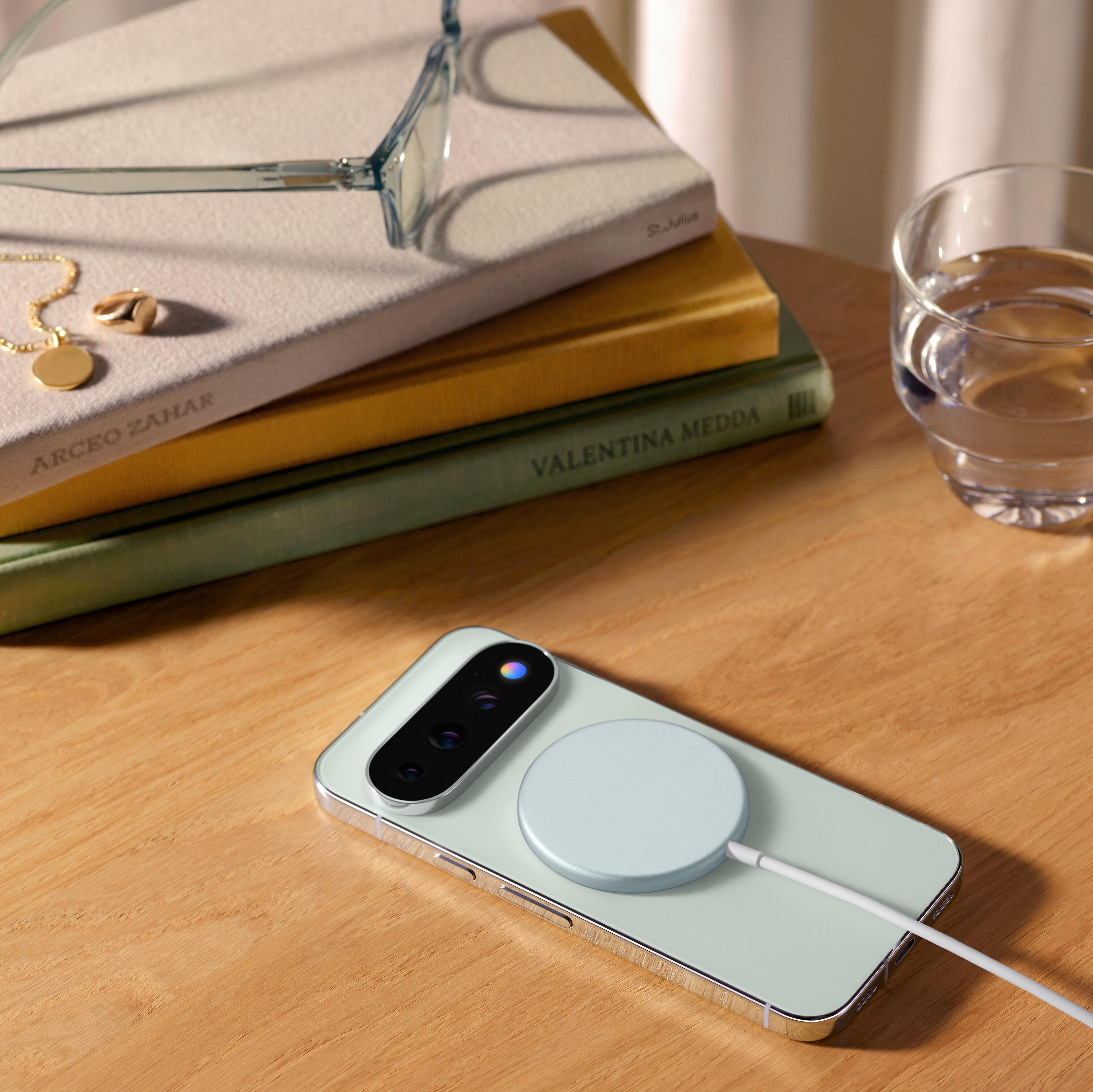 Ein Smartphone lädt auf einem Holztisch neben gestapelten Büchern, Brillen und einem Glas Wasser.