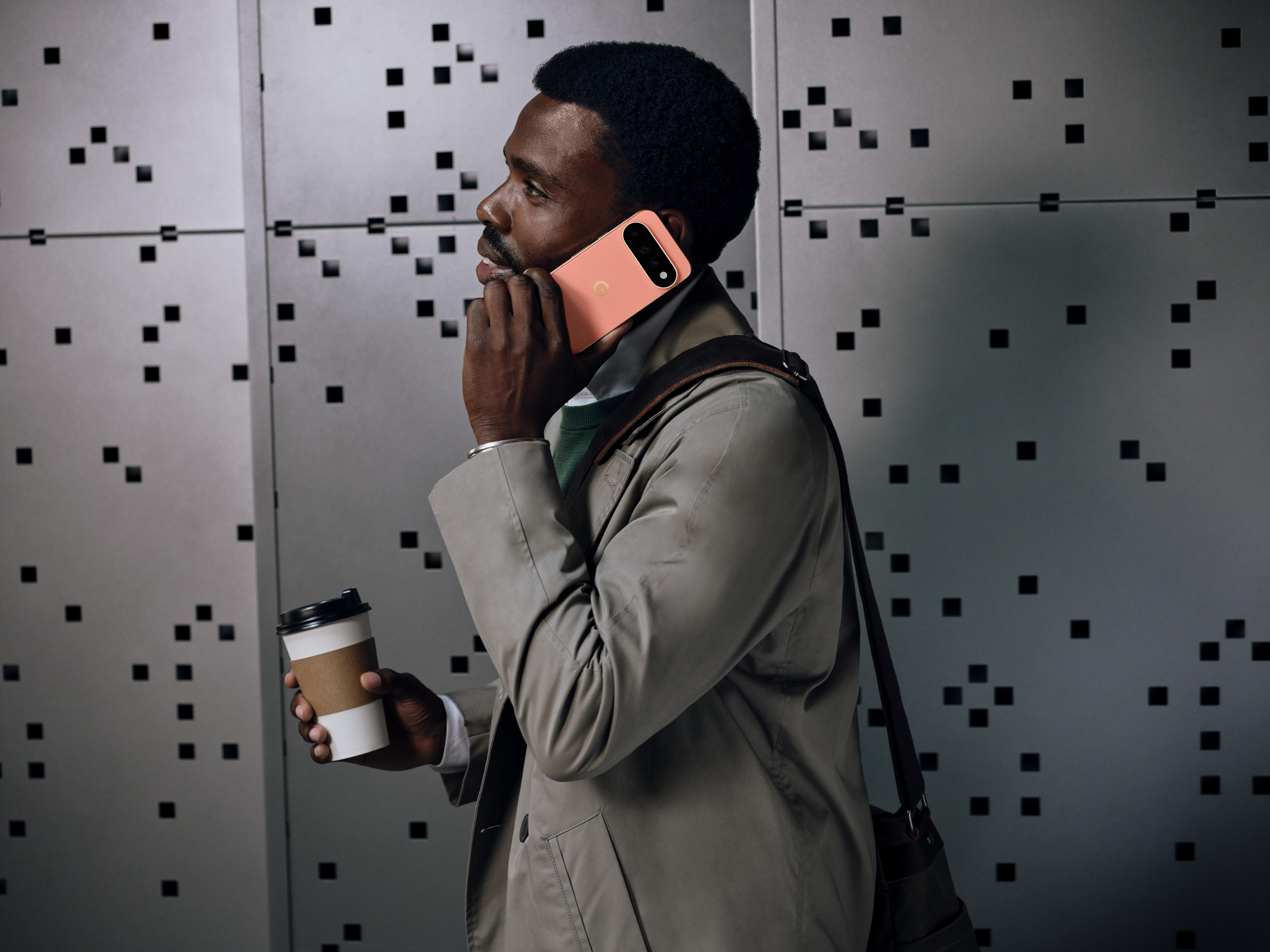 Ein Mann im grauen Mantel telefoniert mit einem rosa Smartphone und hält einen Kaffeebecher vor einer Metallwand.