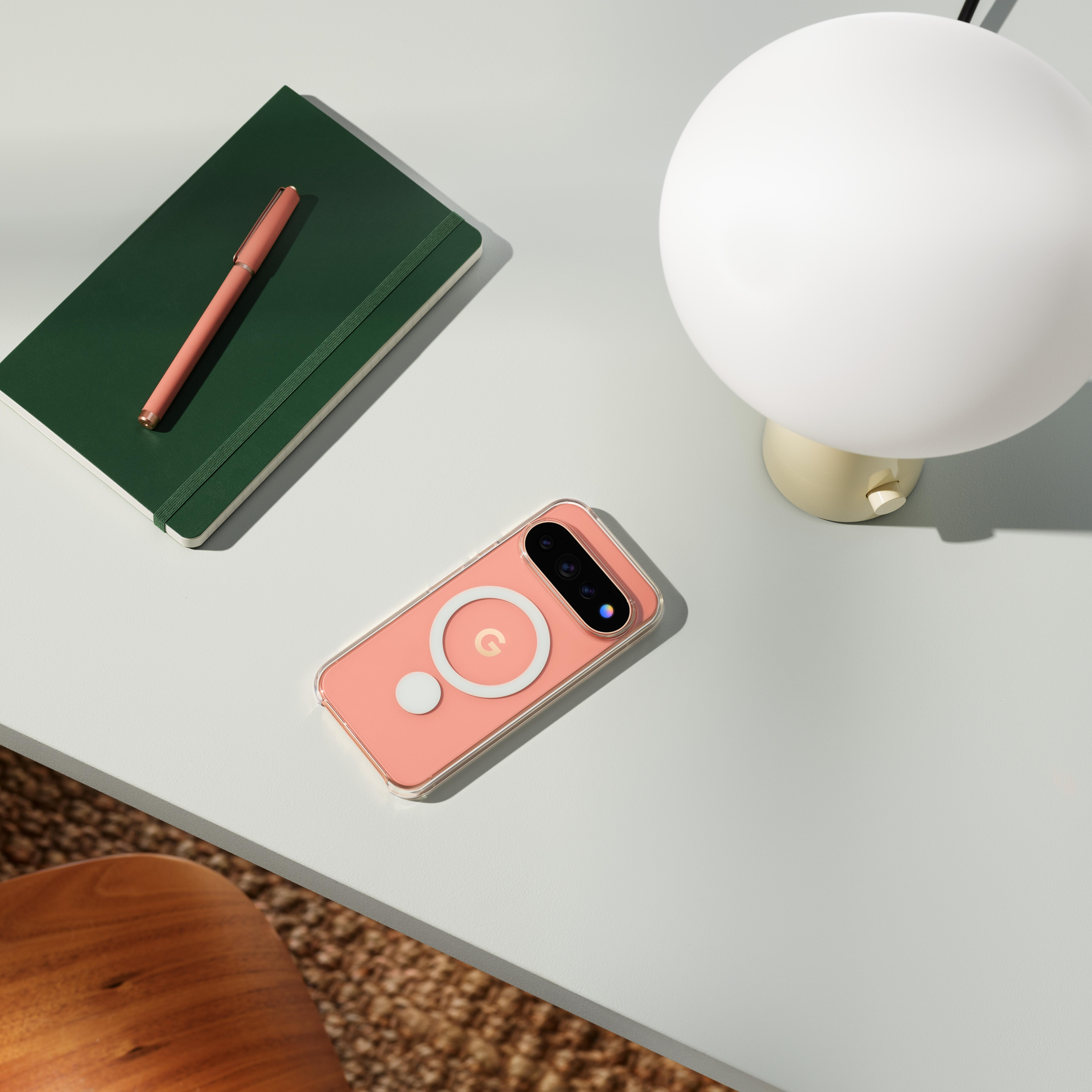 Ein rosafarbenes Smartphone, ein grünes Notizbuch mit Stift und eine weiße Lampe liegen auf einem Schreibtisch.