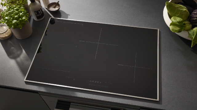 Placa de inducción negra con marco de acero inoxidable en encimera oscura, rodeada de hierbas y verduras frescas.