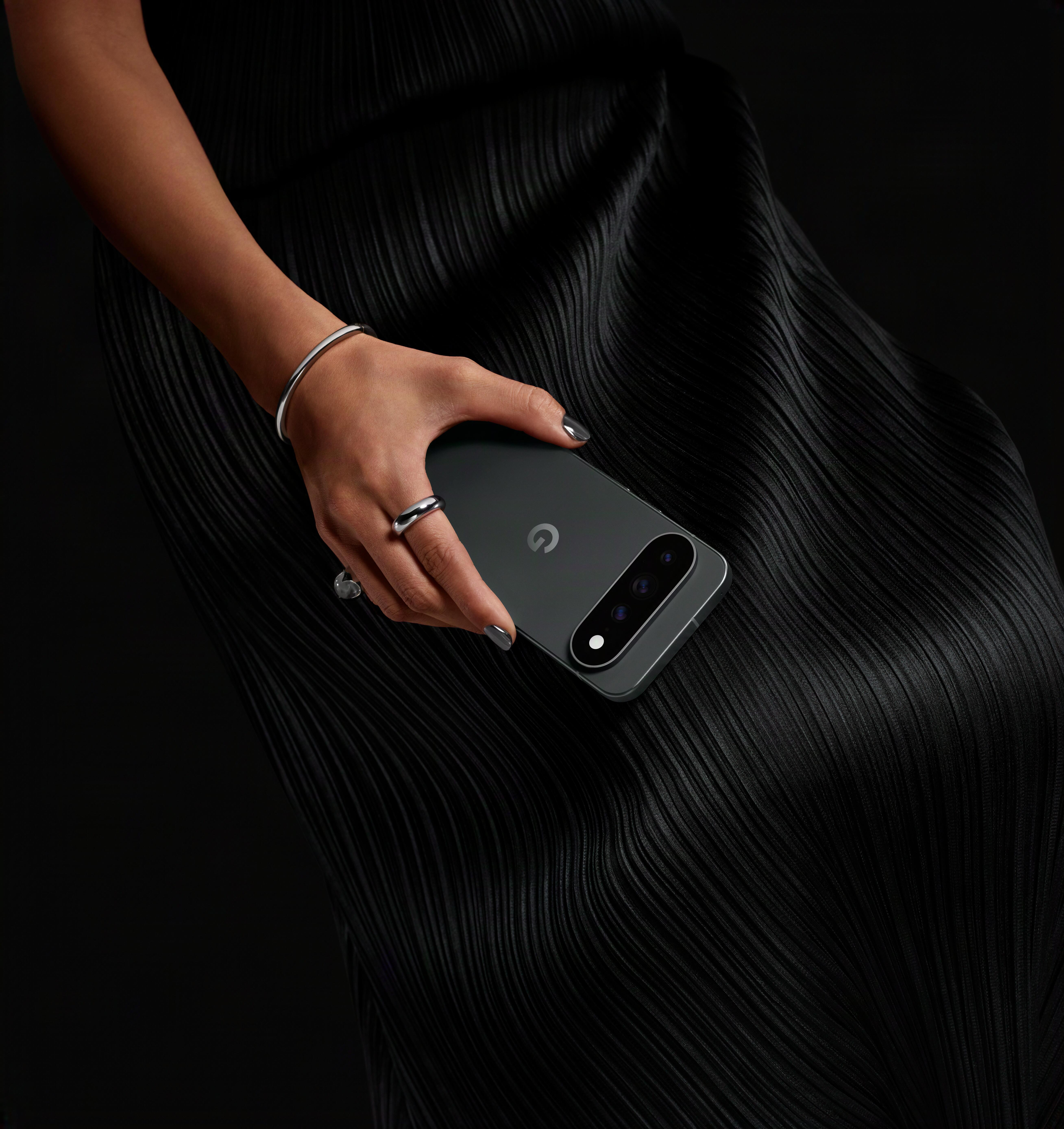 Eine Person mit schwarzem Plisseekleid hält ein graues Smartphone mit Kamera auf der Rückseite vor schwarzem Hintergrund.