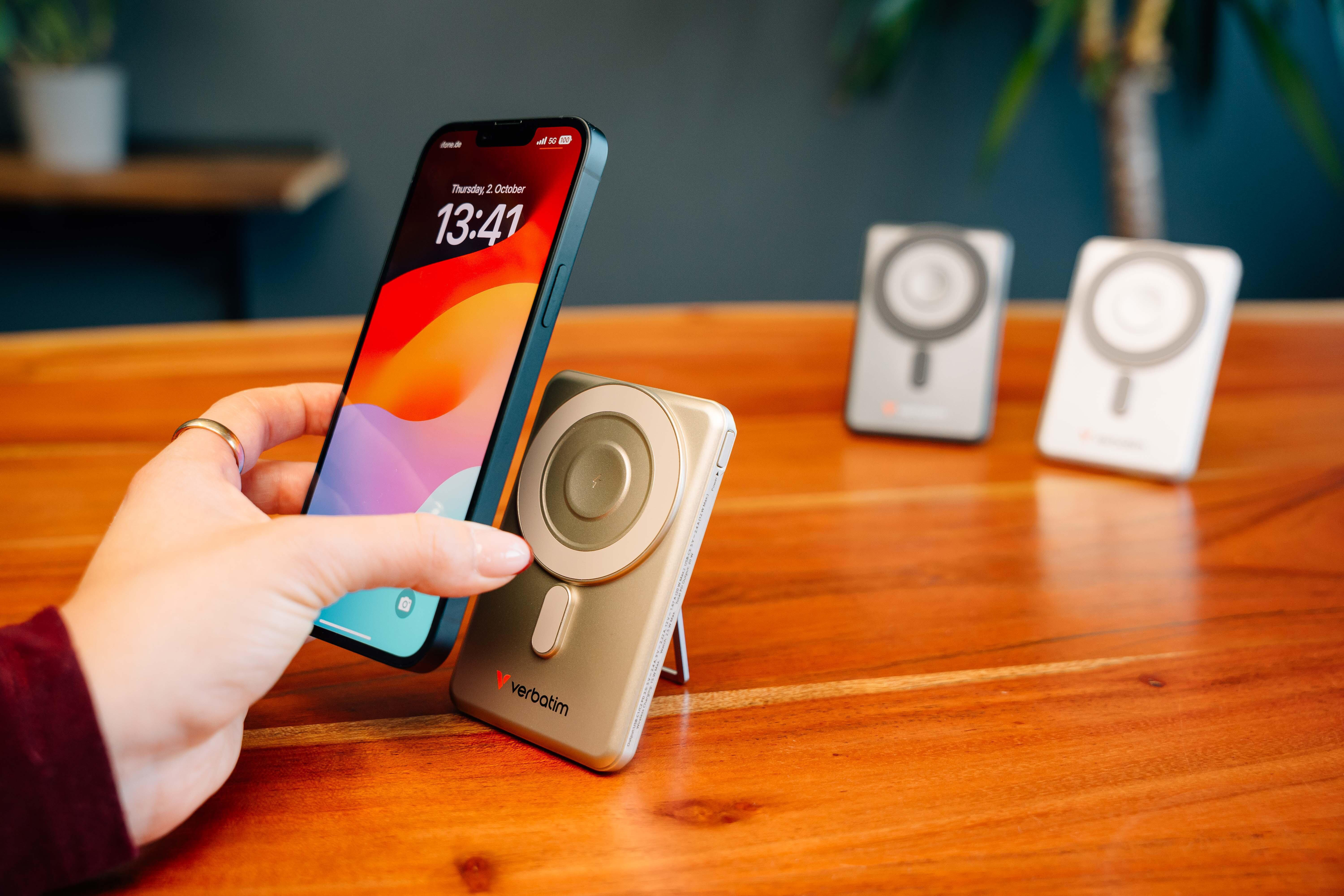 Hand hält Smartphone über eine goldene Verbatim Powerbank auf einem Holztisch, zwei weitere Powerbanks im Hintergrund.