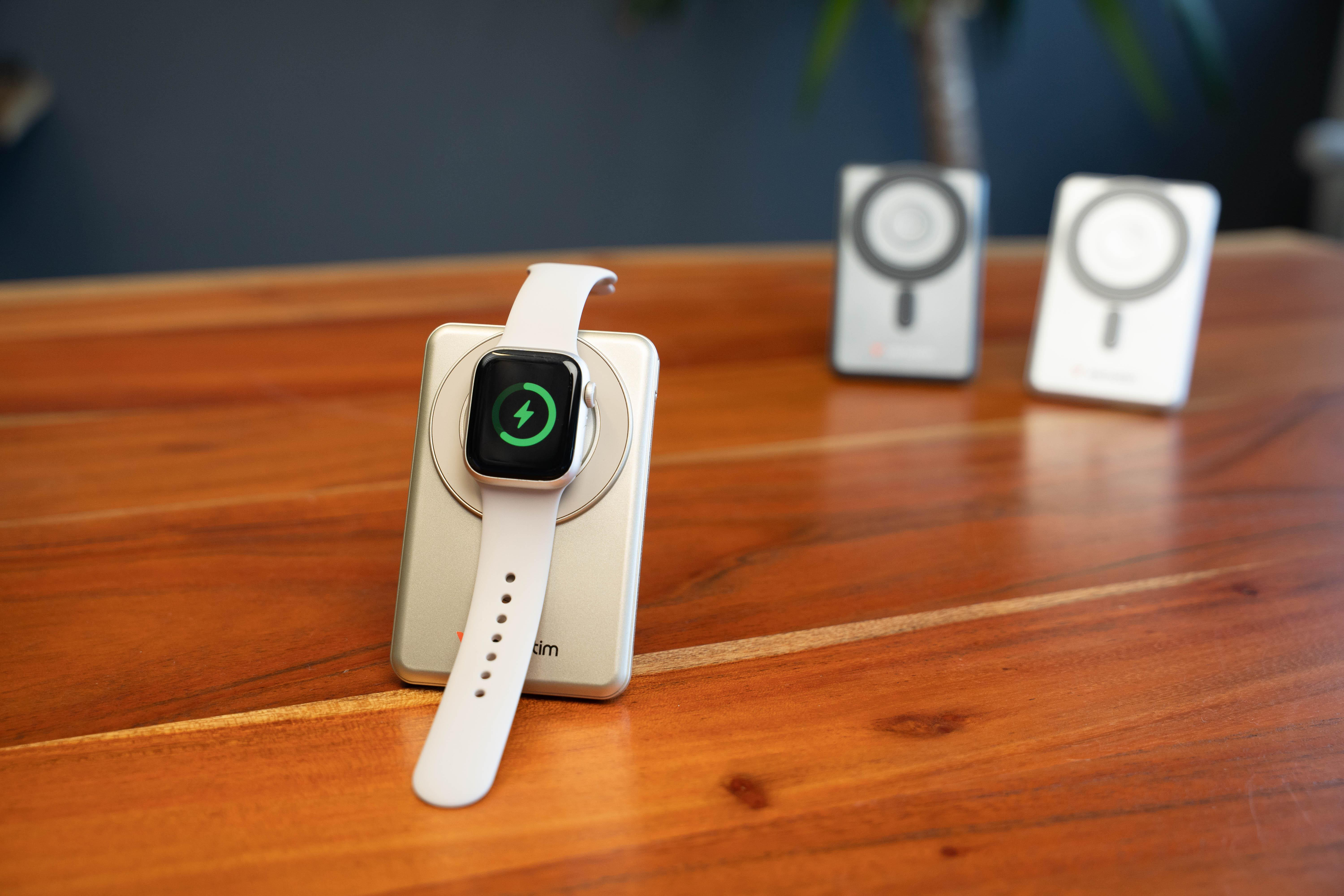 Eine Smartwatch mit weißem Armband lädt auf einem Metallständer auf einem Holztisch, zwei weitere Ständer im Hintergrund.