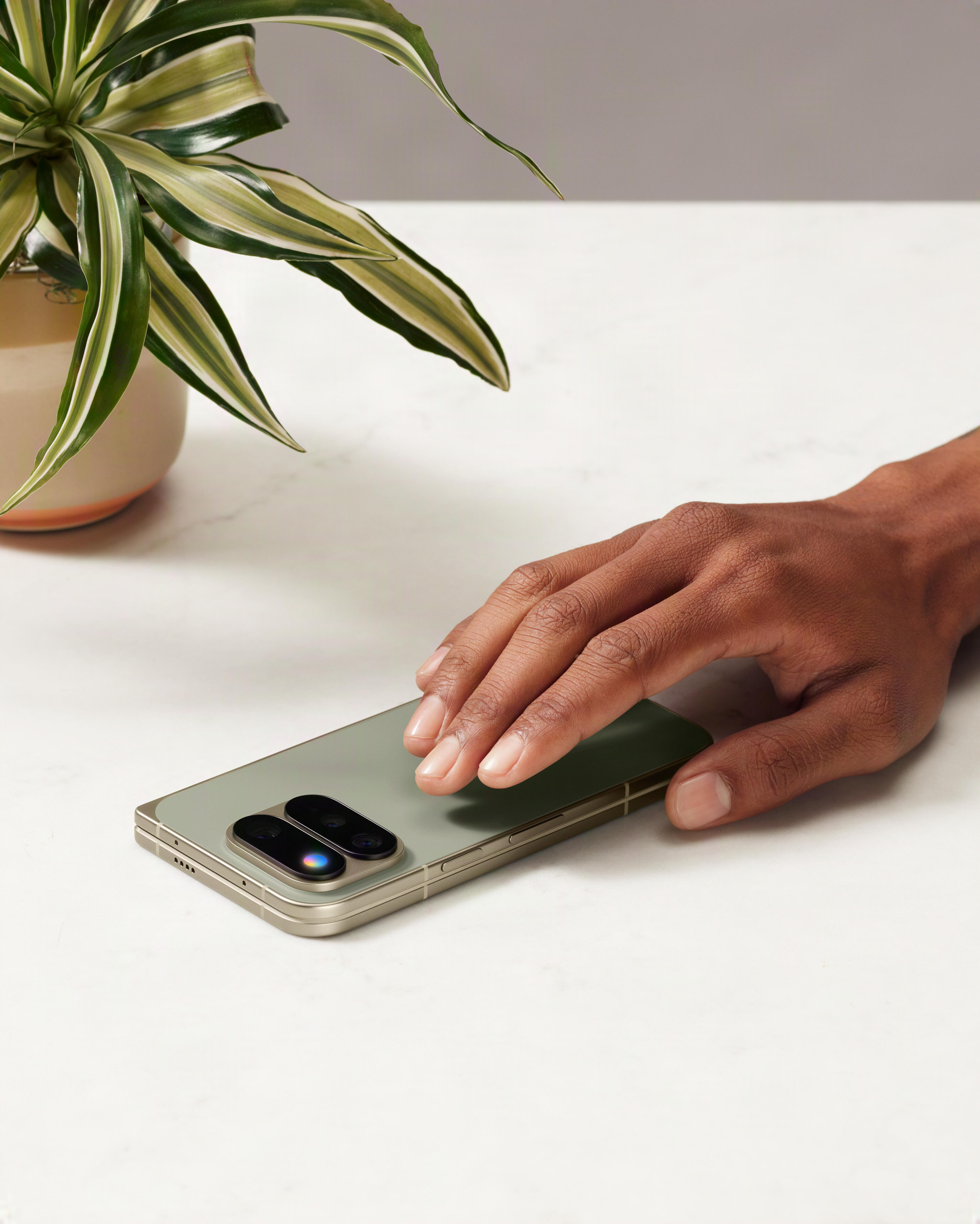 Eine Hand berührt ein grünes Smartphone auf einem weißen Tisch neben einer Topfpflanze.