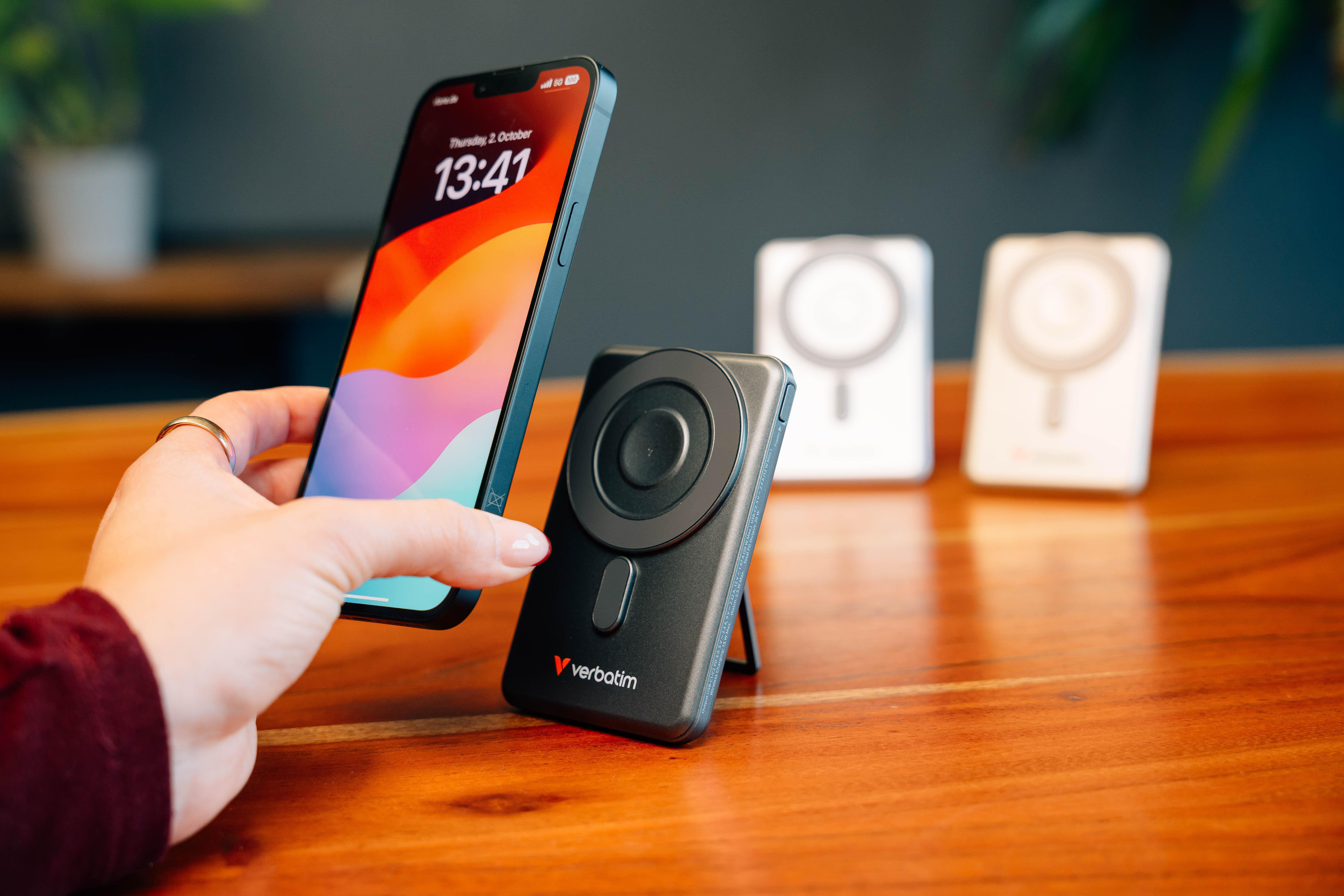 Eine Hand hält ein Smartphone neben ein schwarzes Verbatim kabelloses Ladegerät auf einem Holz Tisch, zwei im Hintergrund.