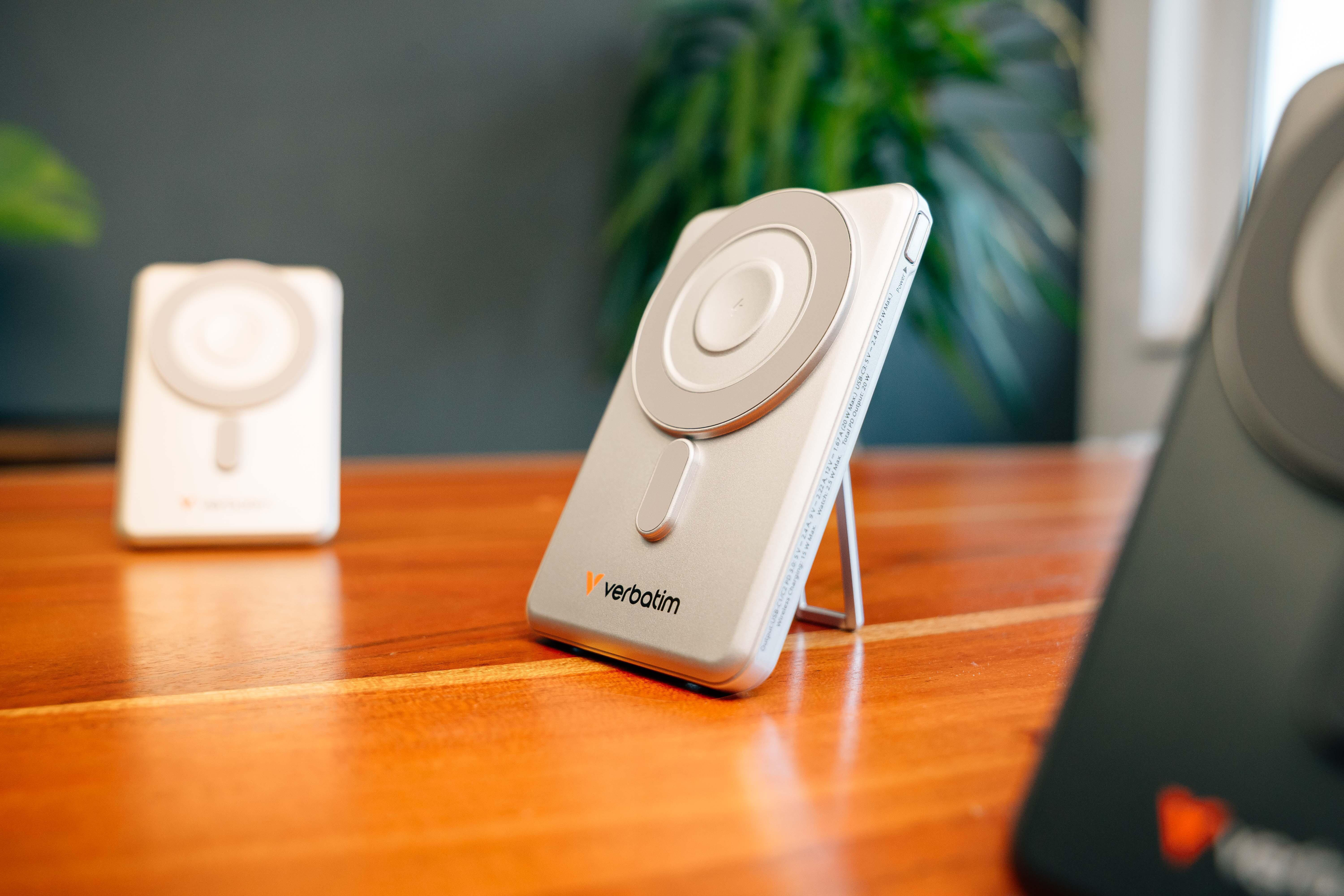 Eine silberne Verbatim Wireless Power Bank steht auf einem Holztisch, im Hintergrund ist eine grüne Pflanze zu sehen.