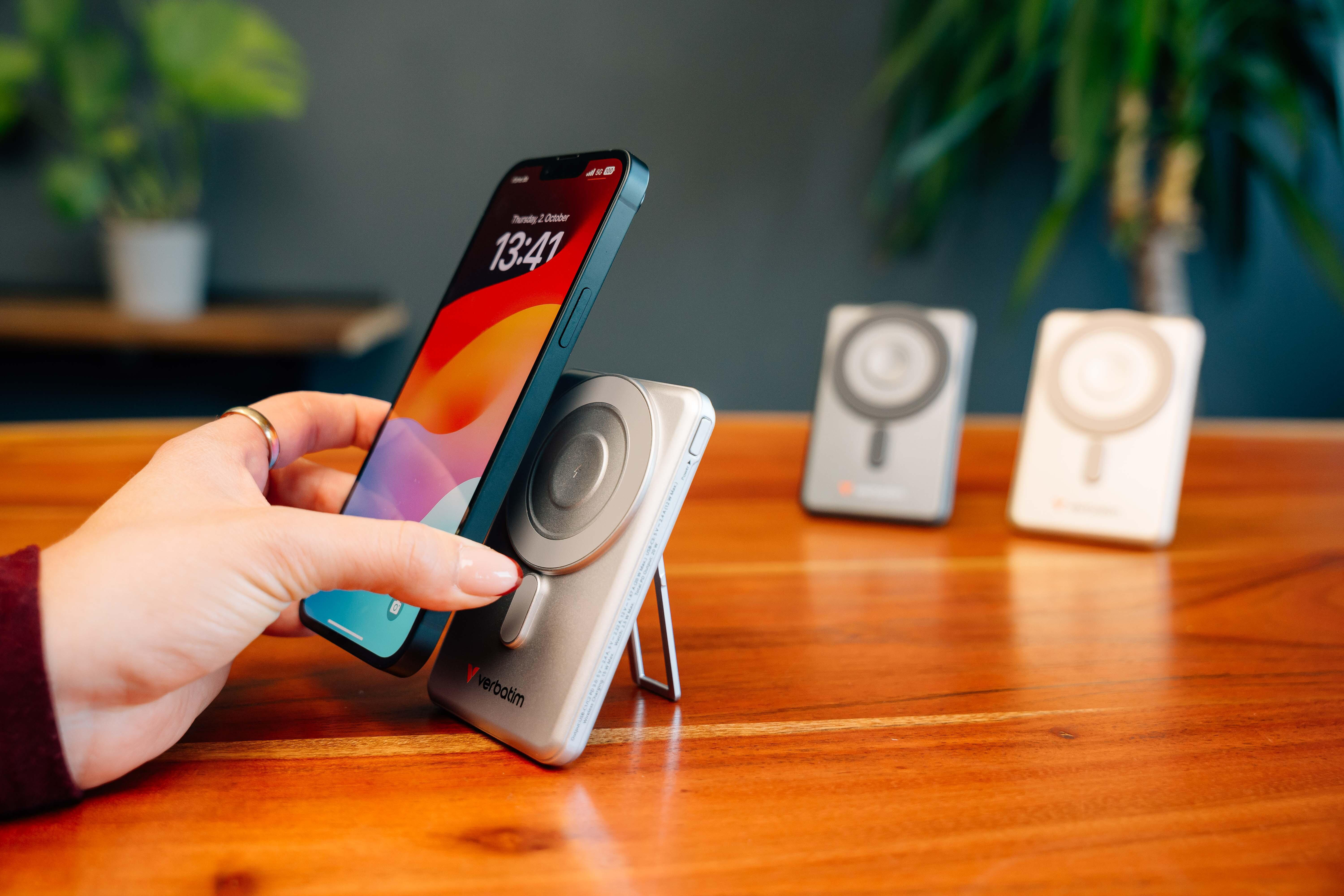 Eine Hand legt ein Smartphone auf eine kabellose Verbatim Ladestation auf einem Holztisch, zwei Ladegeräte im Hintergrund.
