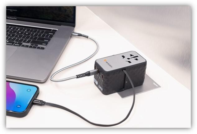 Ein Reiseadapter auf einem Schreibtisch lädt einen Laptop und ein Smartphone mit angeschlossenen Kabeln.