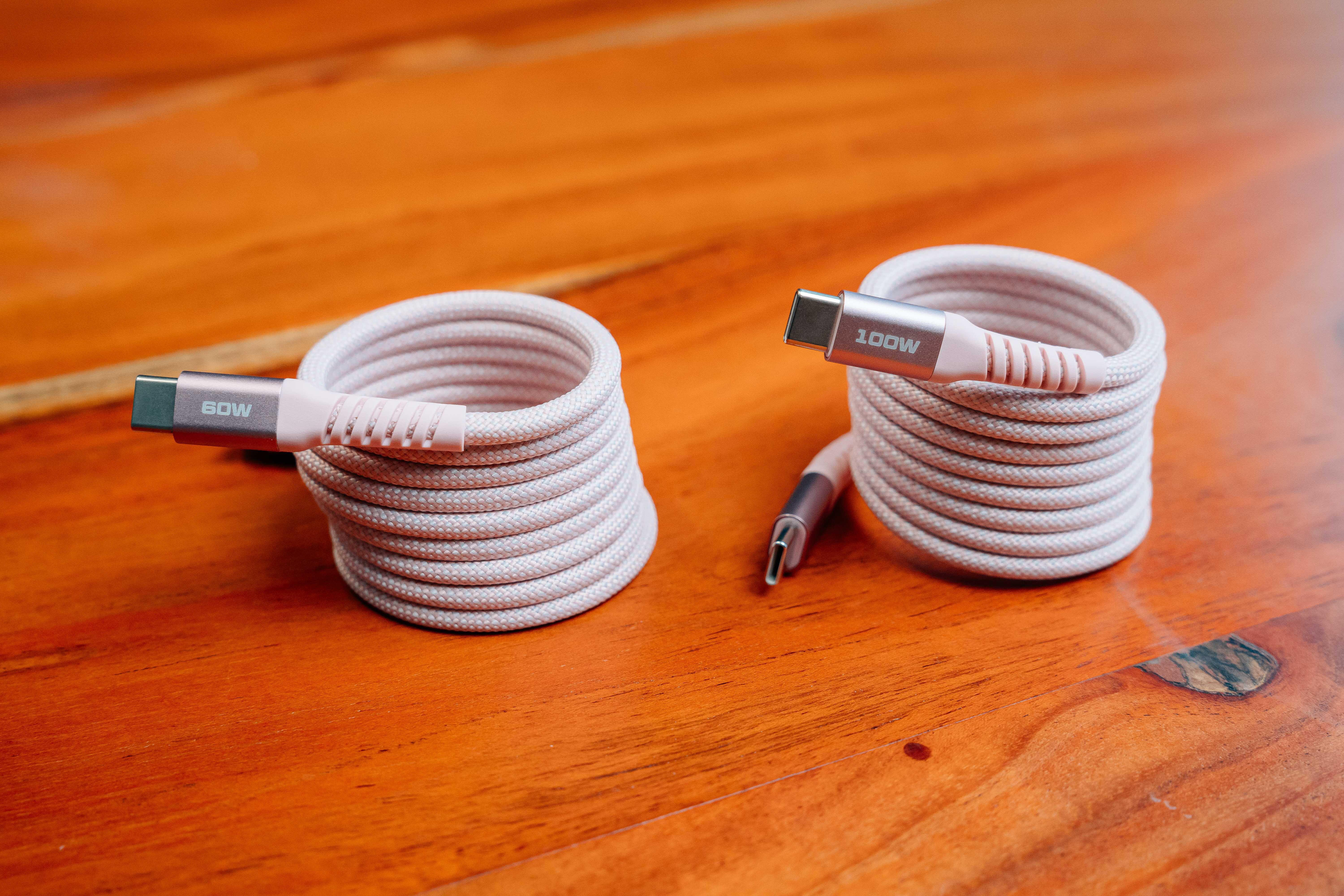 Zwei aufgerollte, rosafarbene USB-C-Kabel, eines mit 60W und eines mit 100W, liegen auf einer Holzoberfläche.