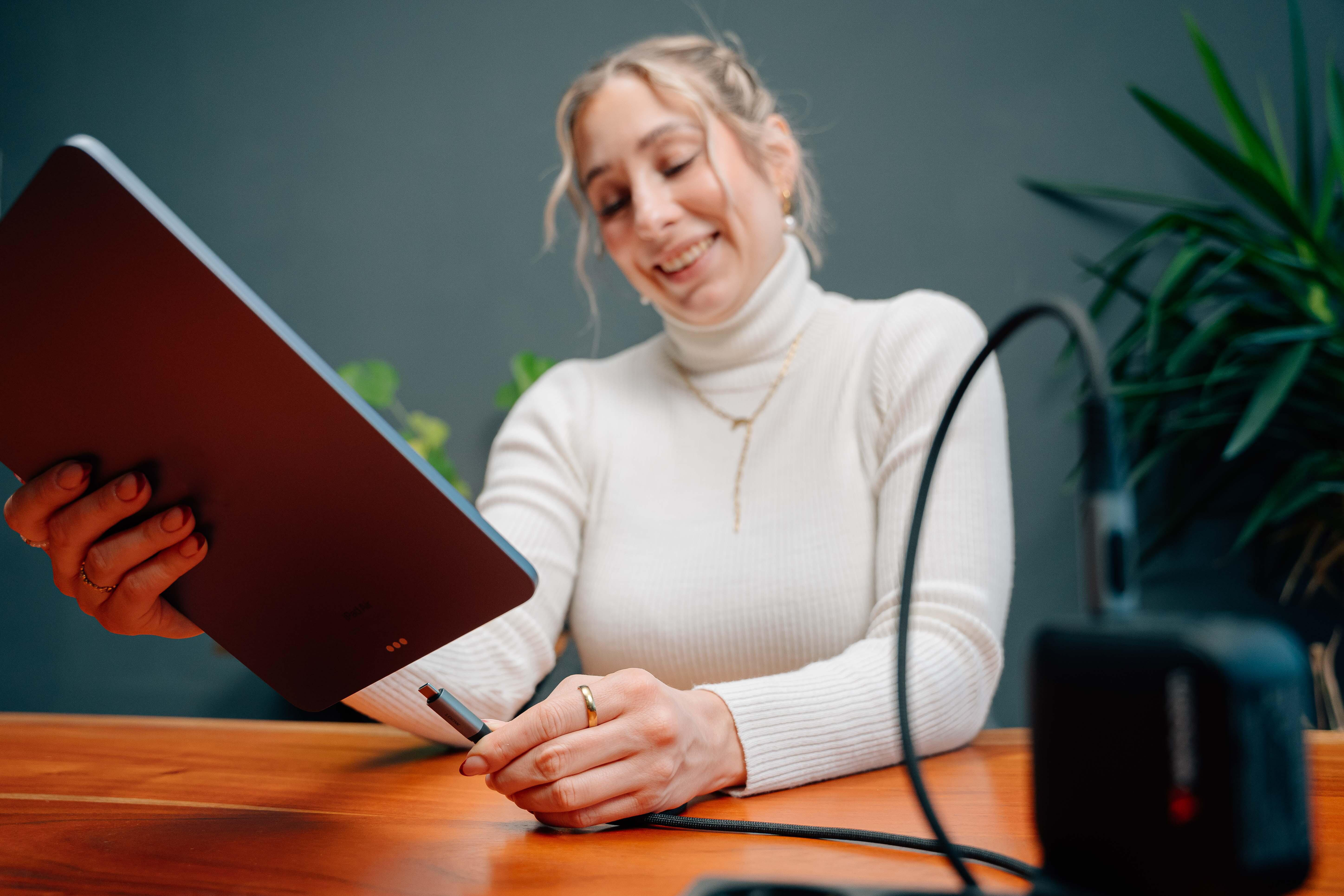 Eine lächelnde Frau im weißen Pullover verbindet ein Ladekabel mit ihrem Tablet an einem Holzschreibtisch.