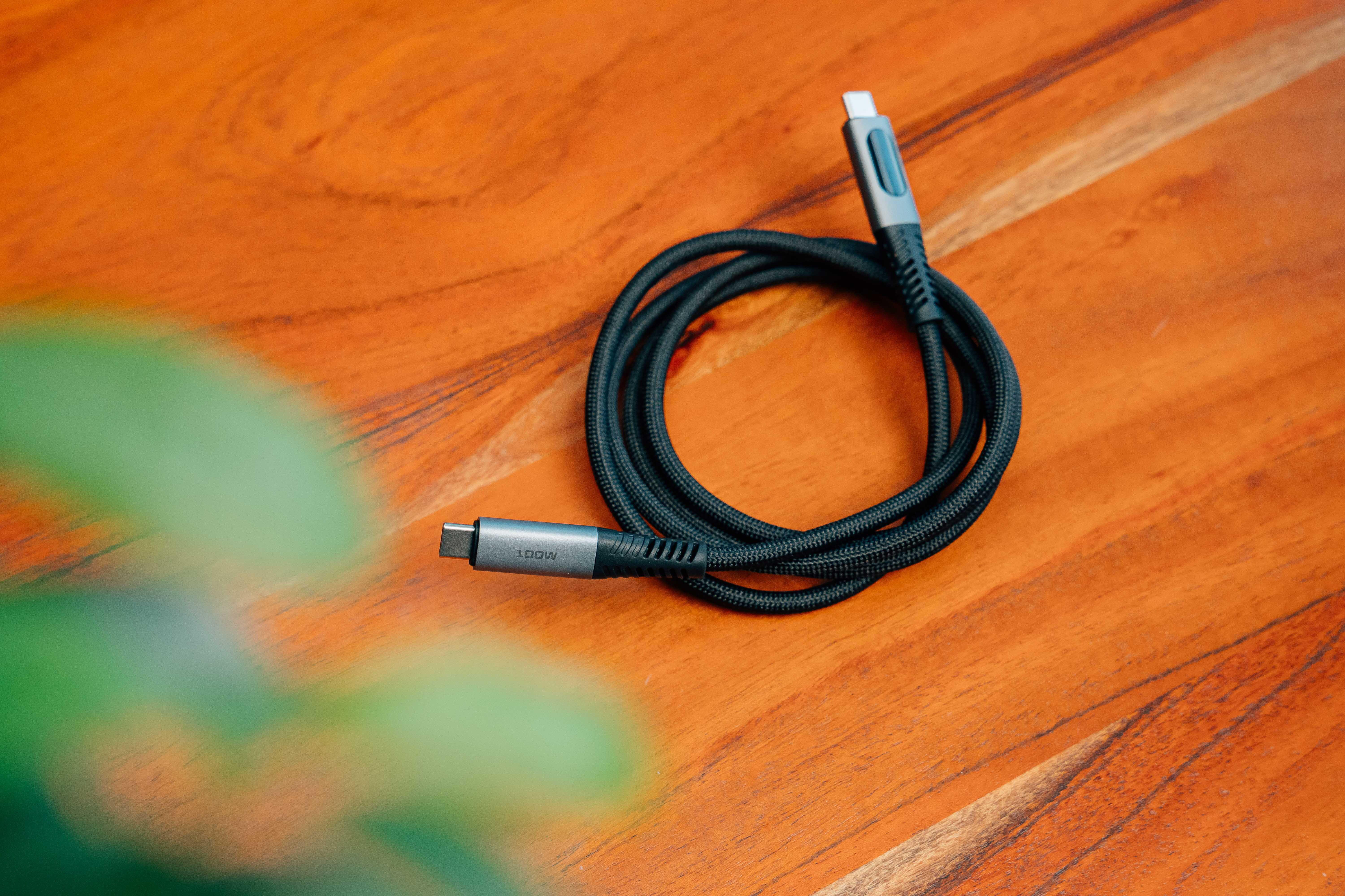 Ein gerolltes schwarzes, geflochtenes USB-C-Kabel mit grauen Steckern auf einem Holztisch, im Vordergrund verschwommene grüne Blätter.