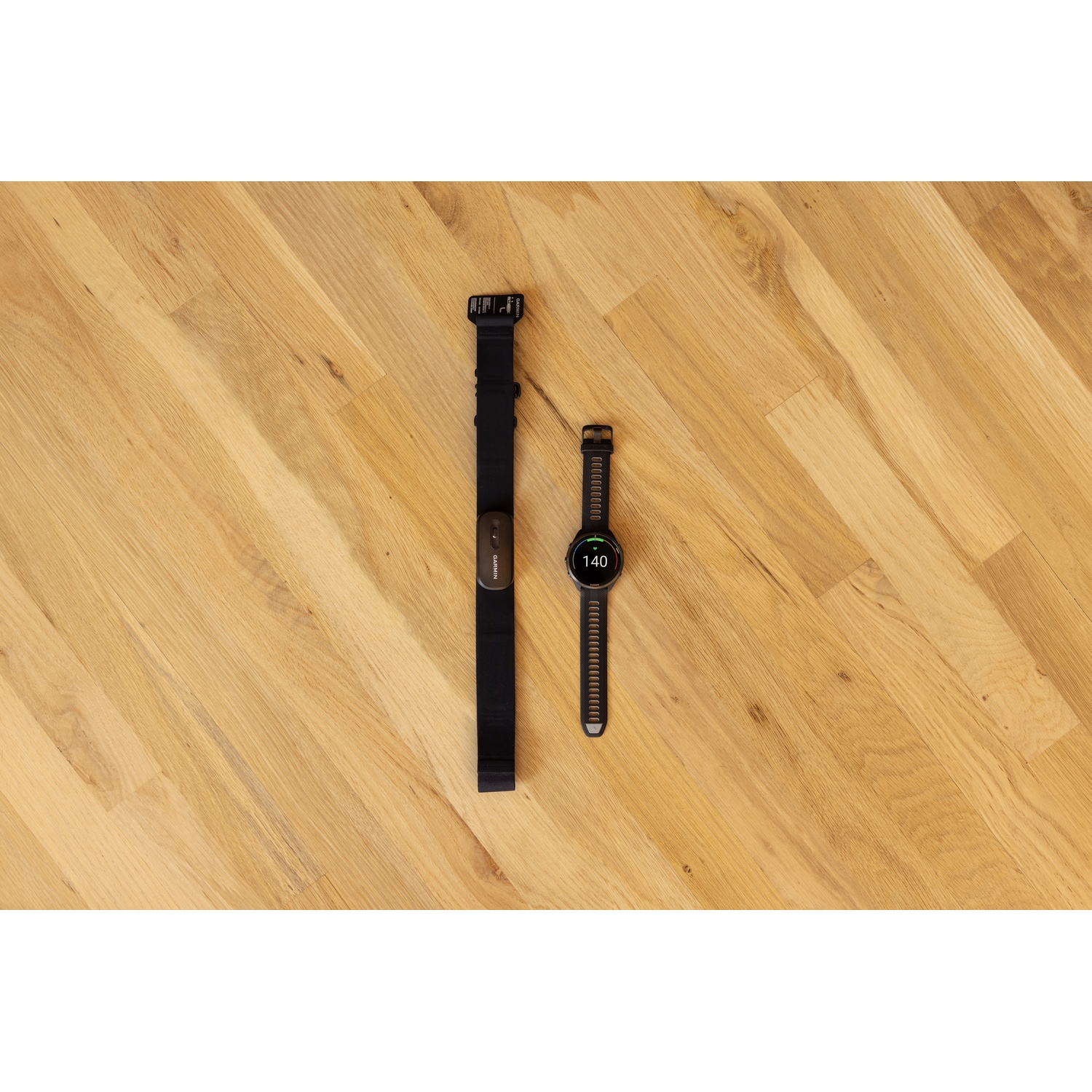 Une sangle de moniteur de fréquence cardiaque Garmin noire et une montre de sport sur un sol en bois clair.