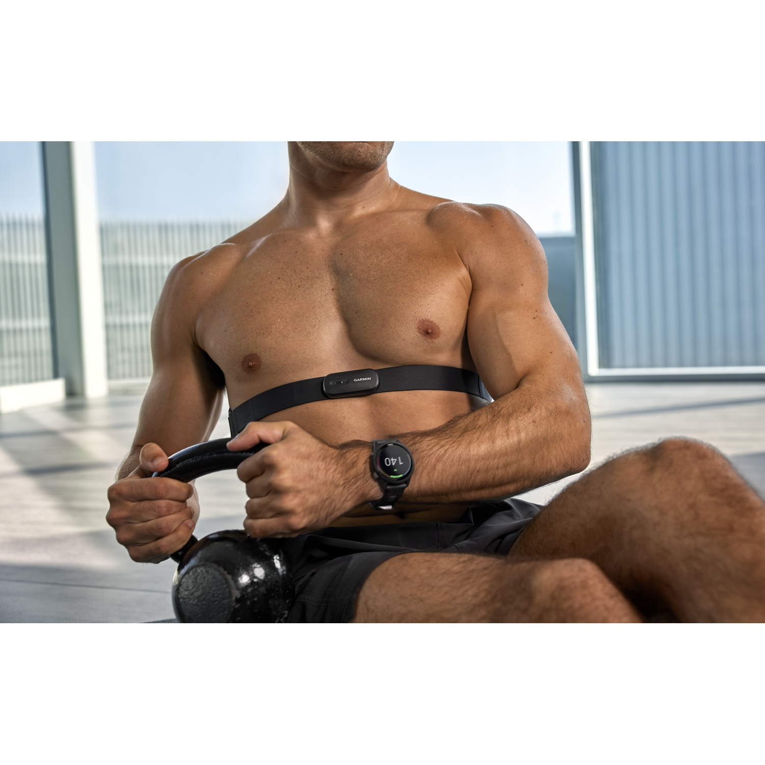 Homme musclé portant une ceinture cardiaque et une montre tient un kettlebell en salle.