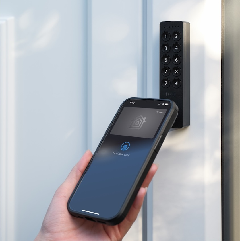 Una mano tiene uno smartphone vicino al tastierino di una serratura intelligente su una porta bianca.