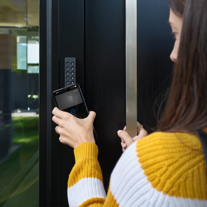 Una donna con un maglione giallo e bianco avvicina uno smartphone alla serratura digitale di una porta.