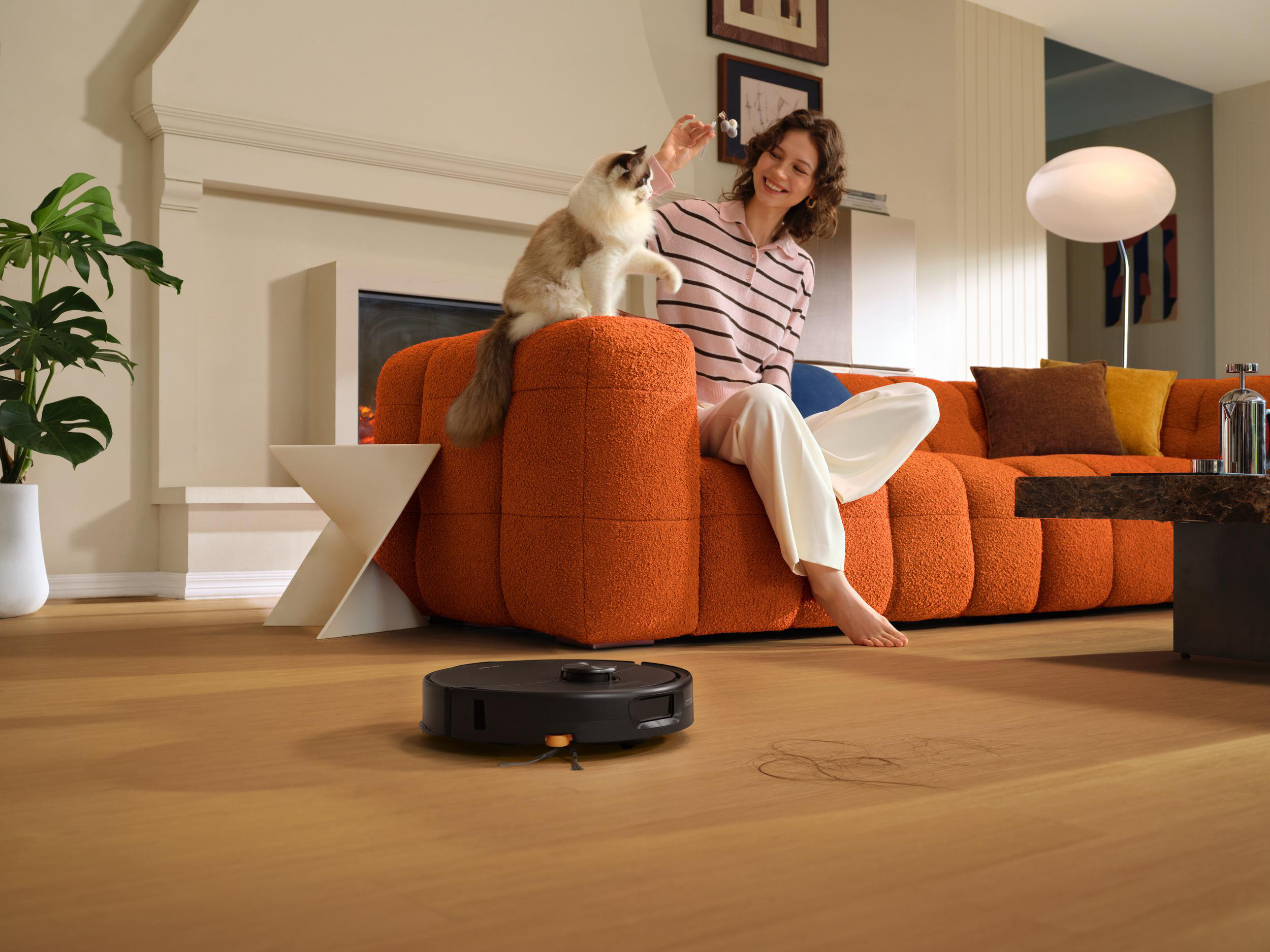 Eine Frau sitzt auf einem orangefarbenen Sofa und spielt mit einer Katze, während ein Saugroboter auf dem Holzfußboden reinigt.