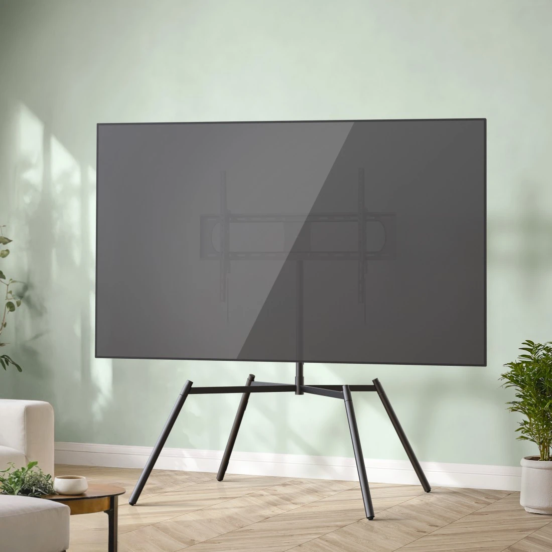 Ein großer Fernseher auf einem schwarzen Ständer steht in einem Raum mit Holzboden und hellgrünen Wänden.