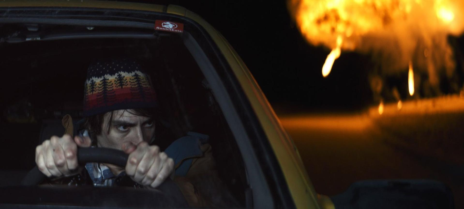 Ein Mann mit Strickmütze greift das Lenkrad im Auto, während draußen ein Feuer explodiert.