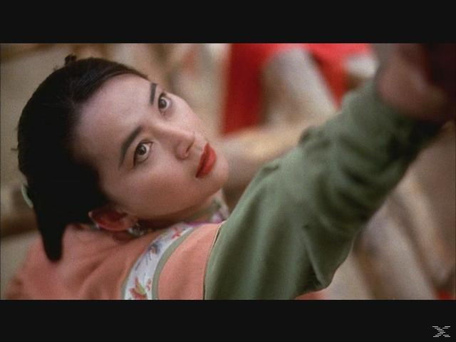 Eine asiatische Frau blickt nach oben, trägt ein grün-oranges Oberteil und hat roten Lippenstift auf.