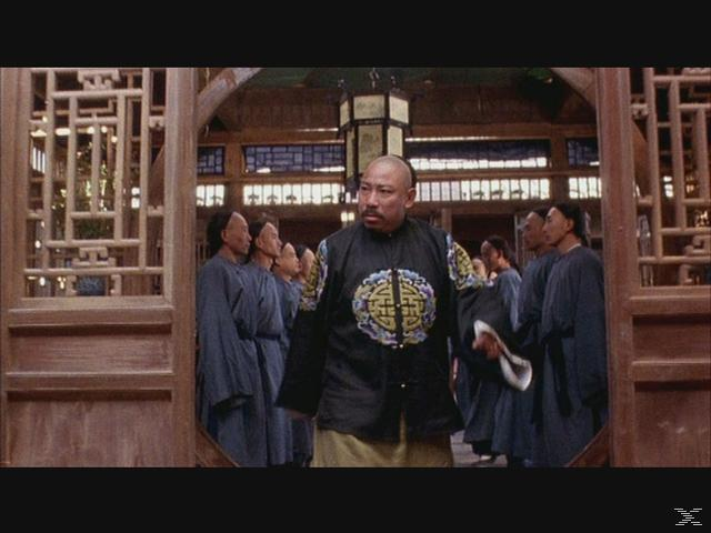 Ein Mann im traditionellen chinesischen Gewand steht in einem holzverkleideten Raum, umgeben von anderen Männern.
