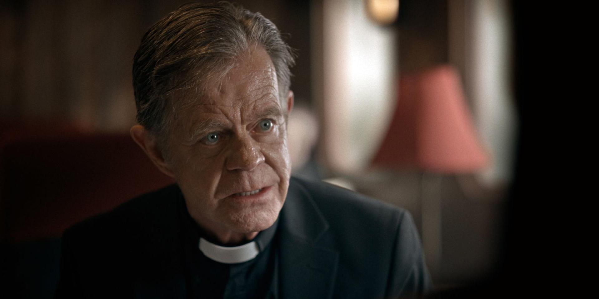 Ein Priester in einem schwarzen Anzug mit Kollarhemd blickt ernst in einem schwach beleuchteten Raum.
