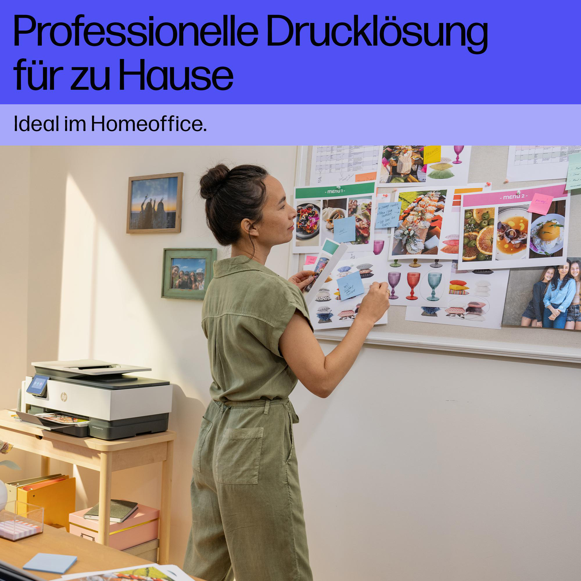 Eine Frau in einem grünen Jumpsuit pinn Fotos an eine große Pinnwand im Homeoffice mit Drucker.