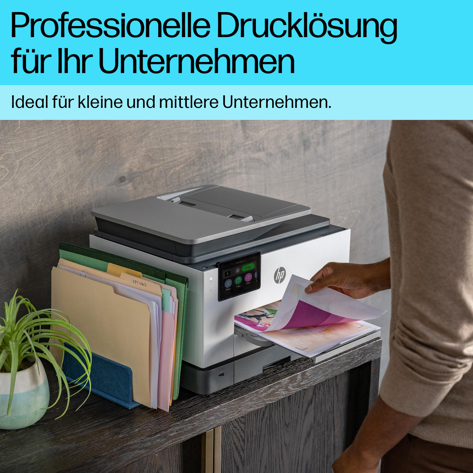 Eine Person greift nach bedrucktem Papier an einem HP-Drucker neben farbigen Aktenordnern und einer Topfpflanze.