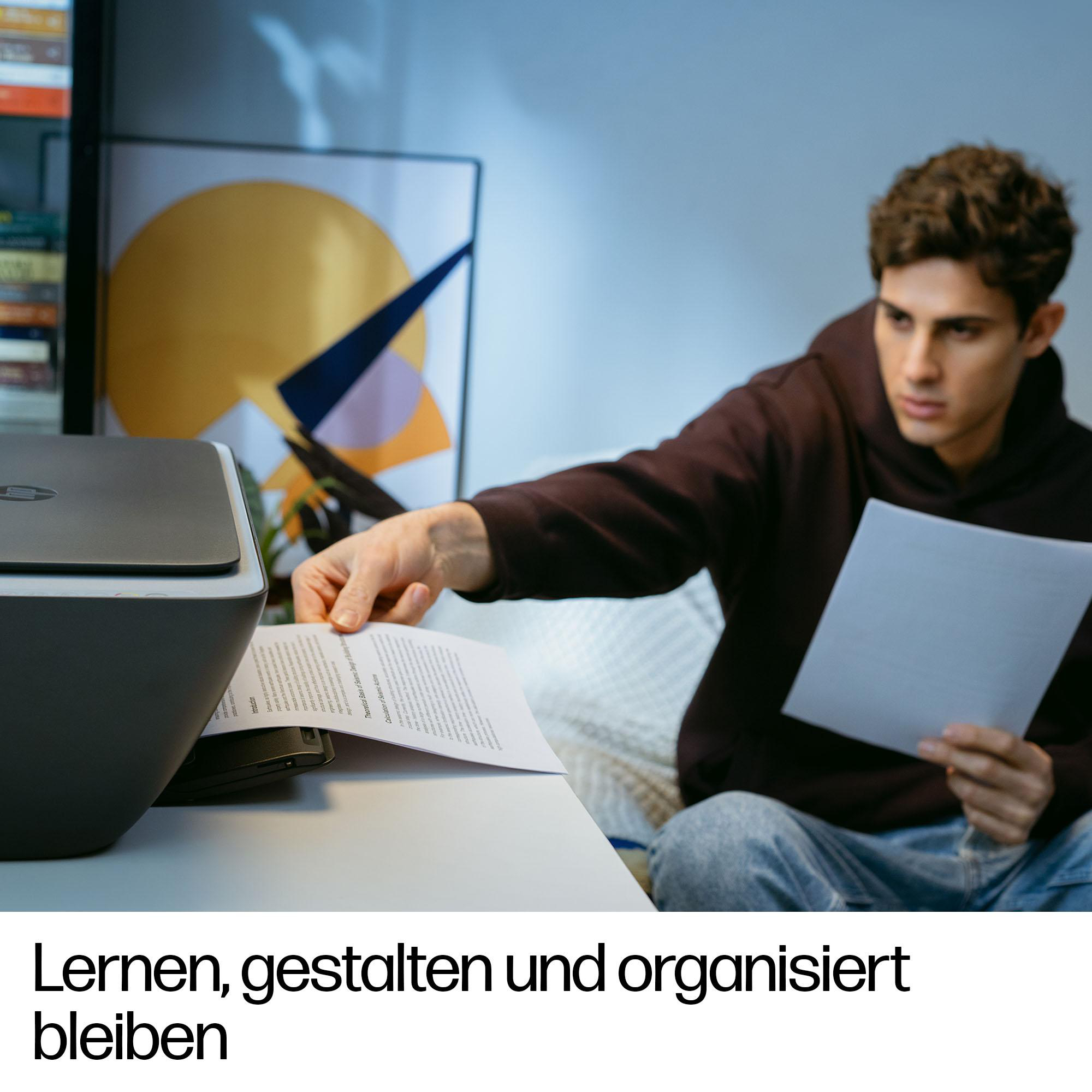 Ein junger Mann im braunen Pullover arbeitet zu Hause am Schreibtisch und druckt Papiere aus.