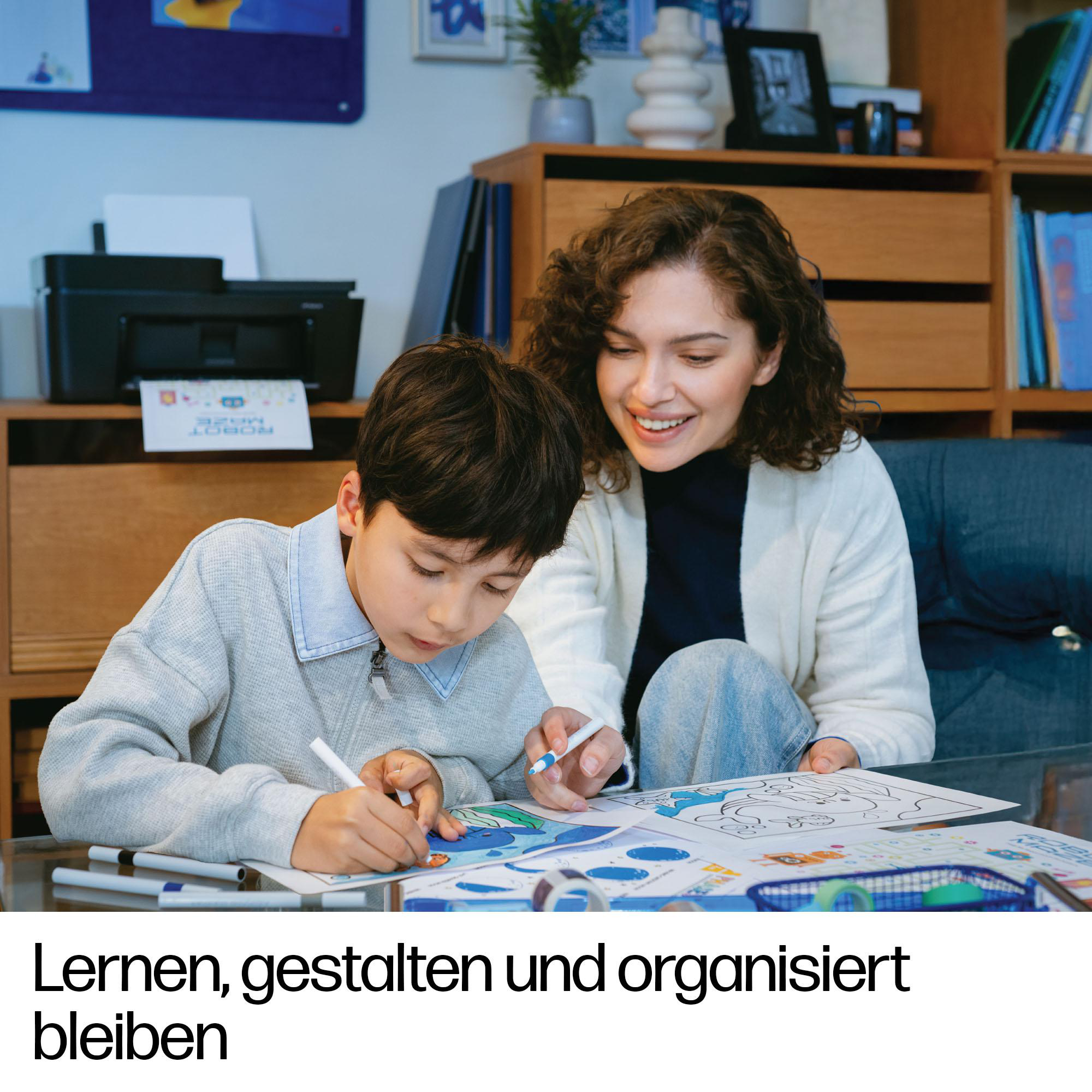 Eine Frau hilft einem Jungen beim Zeichnen an einem Tisch in einem Zimmer mit Drucker und Regalen.