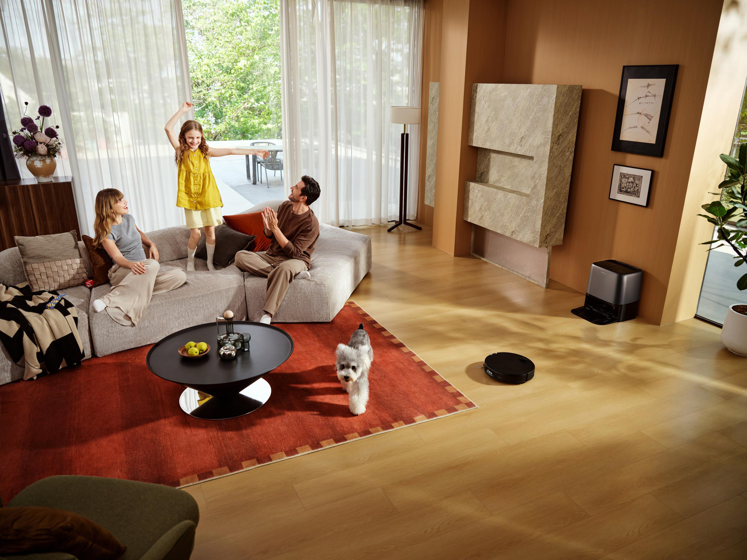 Familie im Wohnzimmer mit Kind, Hund und Roboterstaubsauger auf Holzboden.
