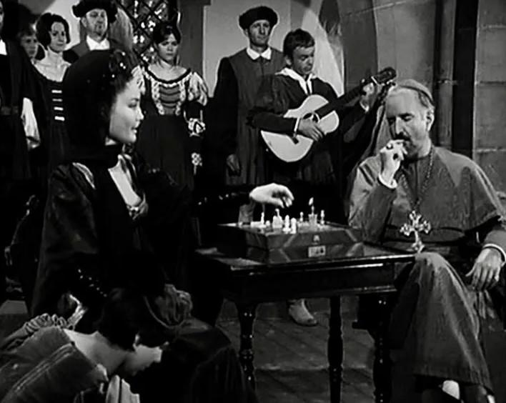 Ein Mann im Priestergewand spielt Schach mit einer Frau, beobachtet von Zuschauern und einem Musiker.