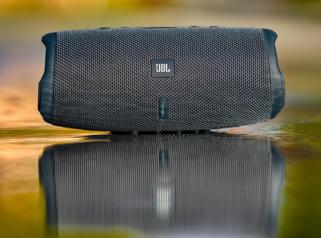 Czarny głośnik Bluetooth JBL spoczywa na mokrej, odbijającej światło powierzchni z rozmytym tłem.