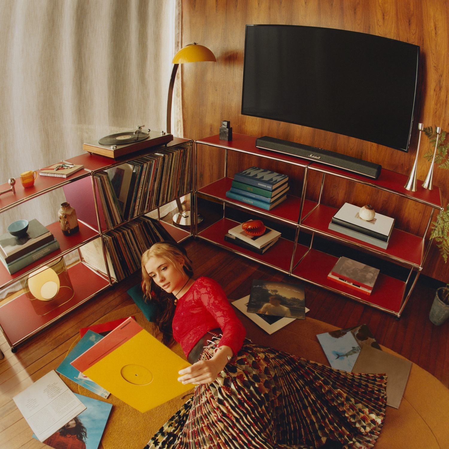 Une femme en haut rouge et jupe à motifs est allongée par terre, regardant des disques vinyles dans un salon rétro.