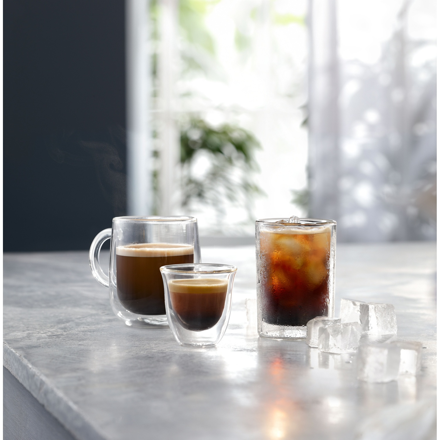 Trois tasses en verre avec café chaud et froid et des glaçons sur une surface claire devant un fond lumineux.
