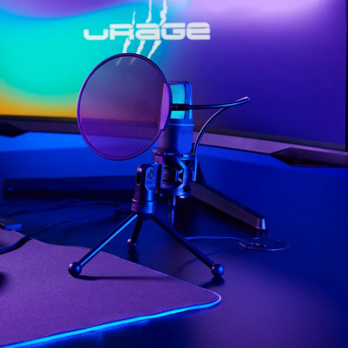 Egy gamer mikrofon popfilterrel egy kis háromlábú állványon, háttérben világító monitorral az asztalon.
