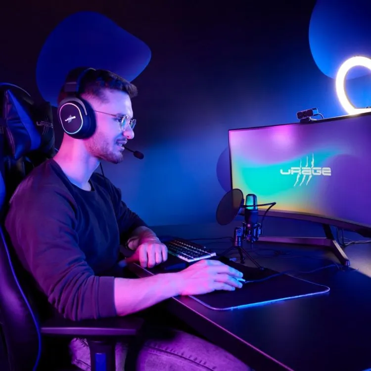 Egy fiatal férfi szemüvegben és fejhallgatóban kék fényben ül egy ívelt monitor előtt, pc-n streamel.