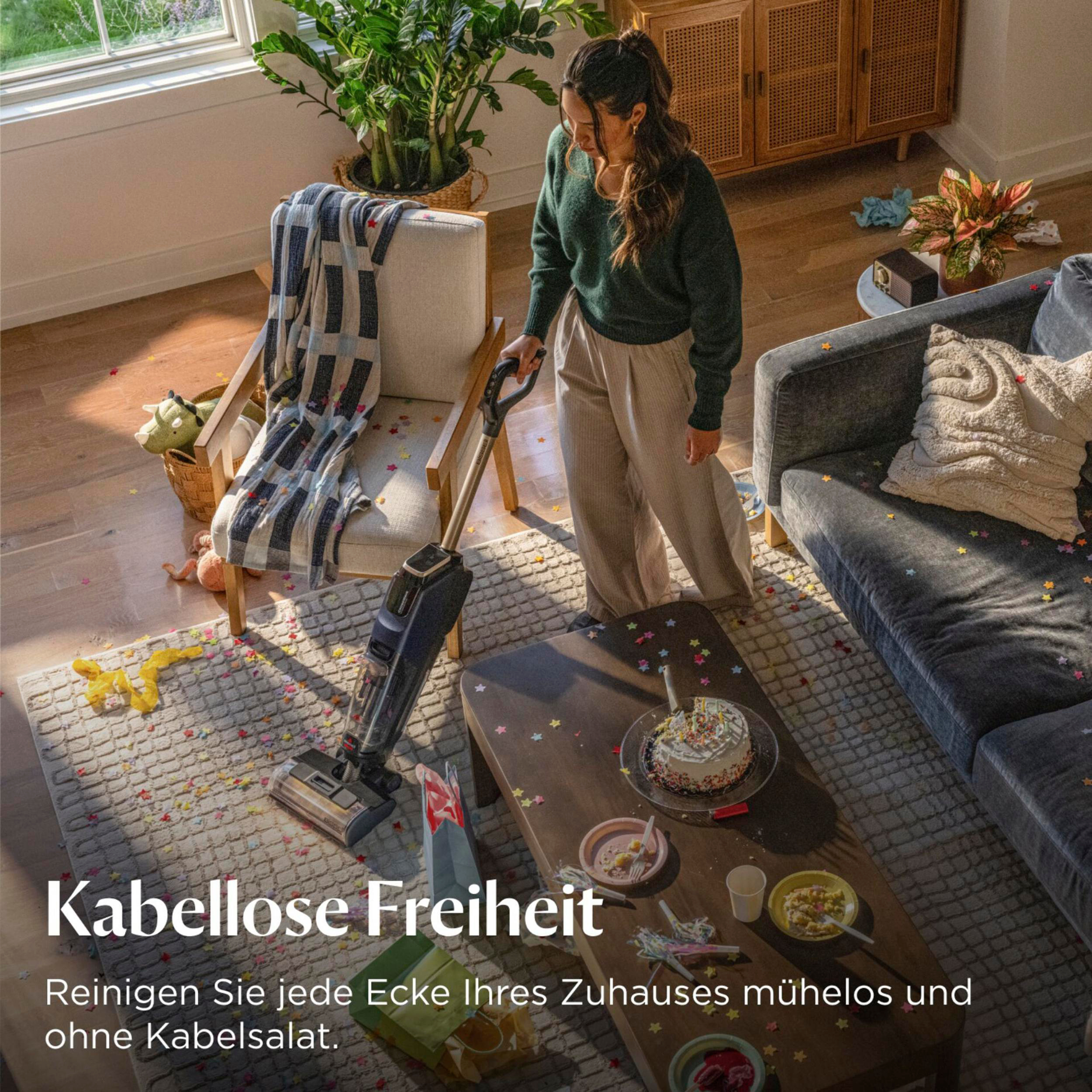 Eine Frau saugt mit einem kabellosen Staubsauger einen konfettibestreuten Wohnzimmerboden neben Sofa und Tisch.
