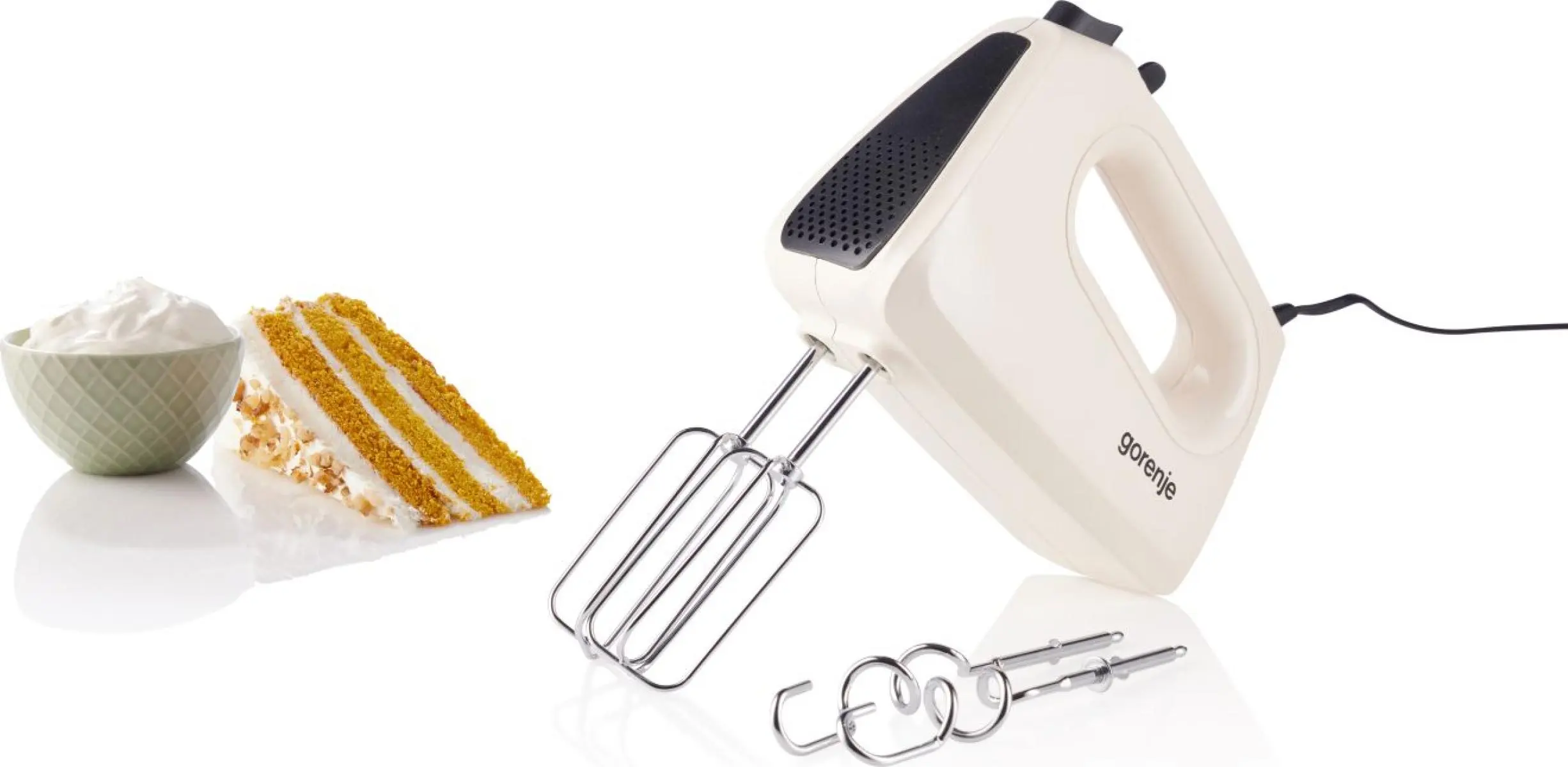 Egy fehér Gorenje kézi mixer, fém keverőszárak, tejszínhab egy tálban és egy szelet sütemény fekete alapon.