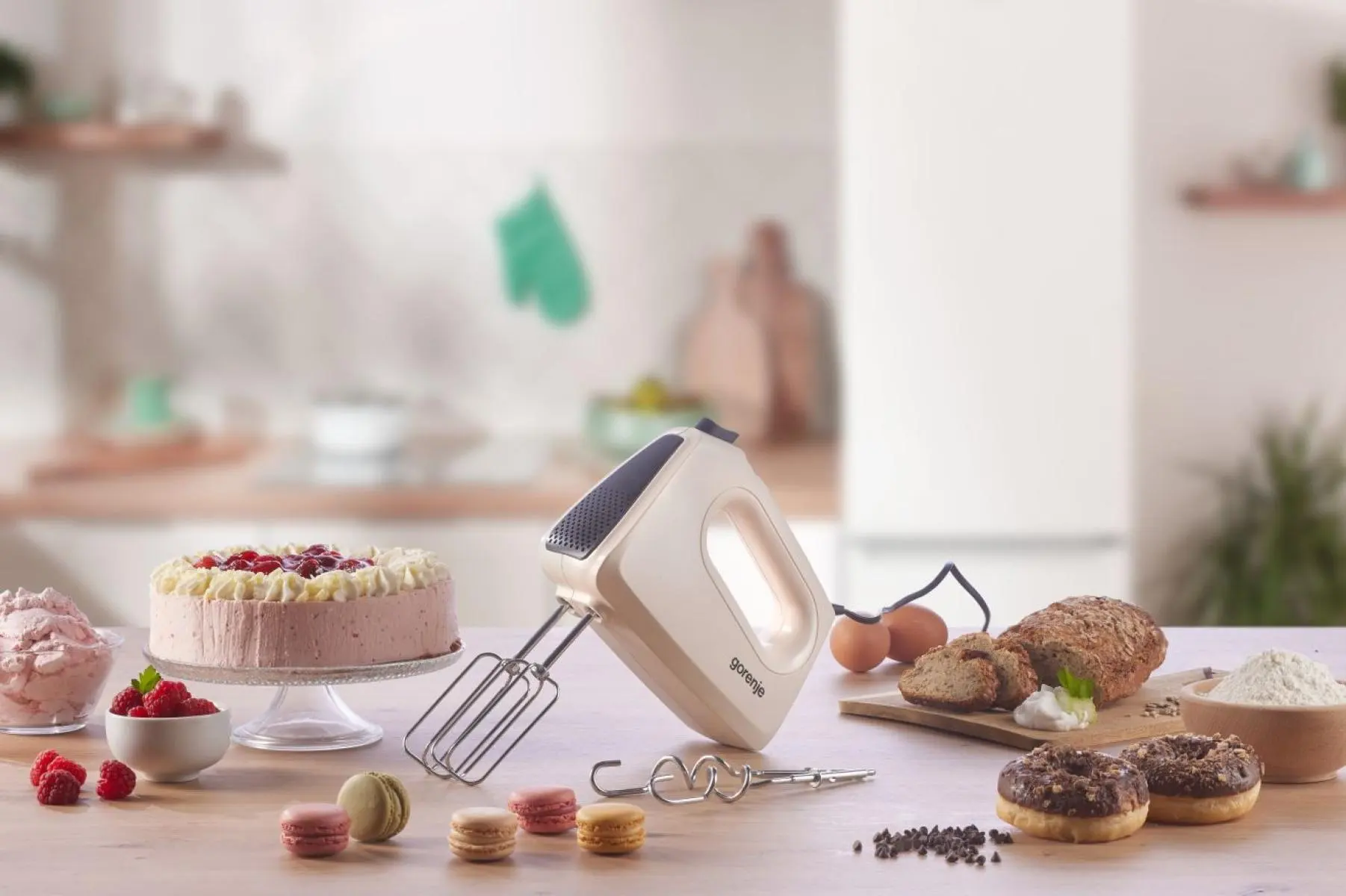 Egy krémszínű kézi mixer áll egy konyhaasztalon, torta, kenyér, fánk és macaronok körében.