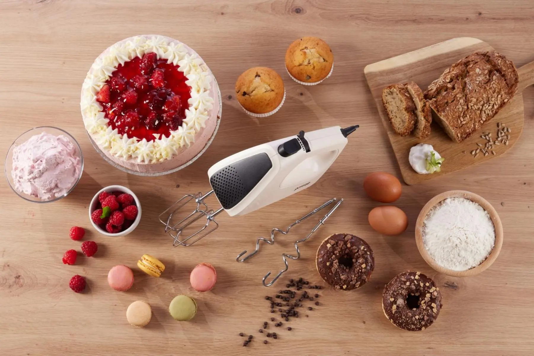 Kézi mixer sütési alapanyagokkal, mint torta, muffin, kenyér, tojás, liszt és édességek egy fa asztalon.