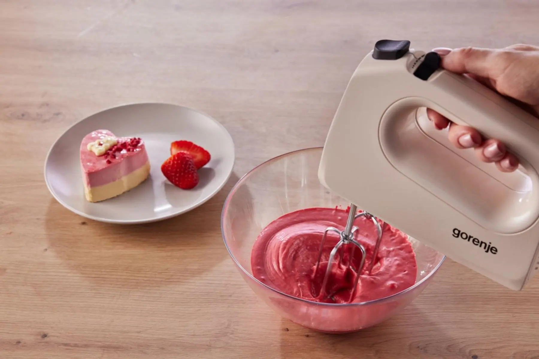 Kézi mixer piros krémet kever egy tálban, mellette szív torta.