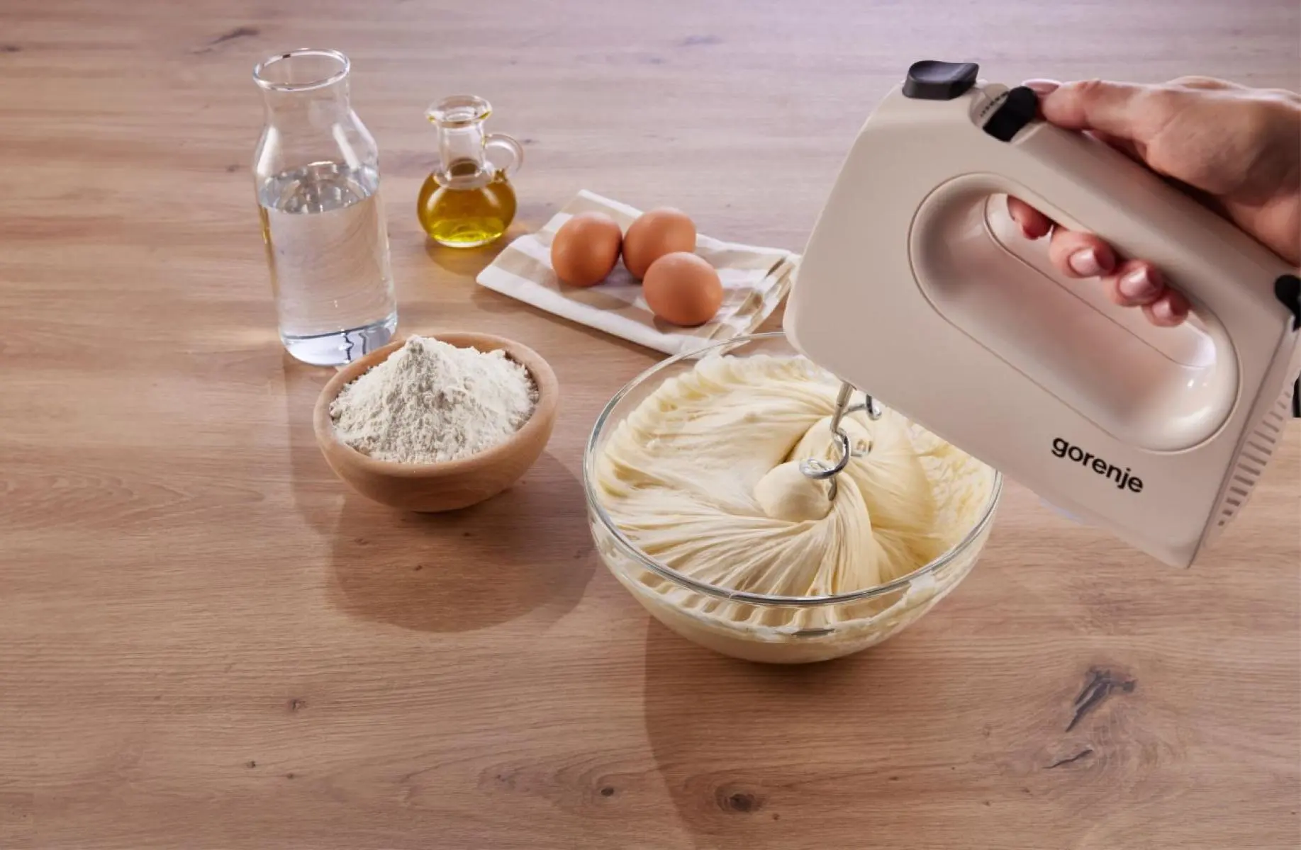 Kézi mixer tésztát kever üvegtálban liszt, tojás, olaj és víz mellett.