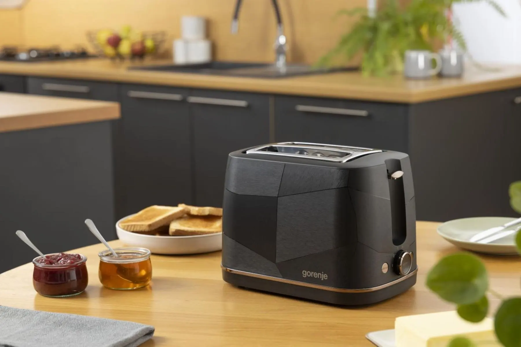 Egy fekete Gorenje kenyérpirító áll egy faasztalon egy modern konyhában pirítóssal és lekvárral.