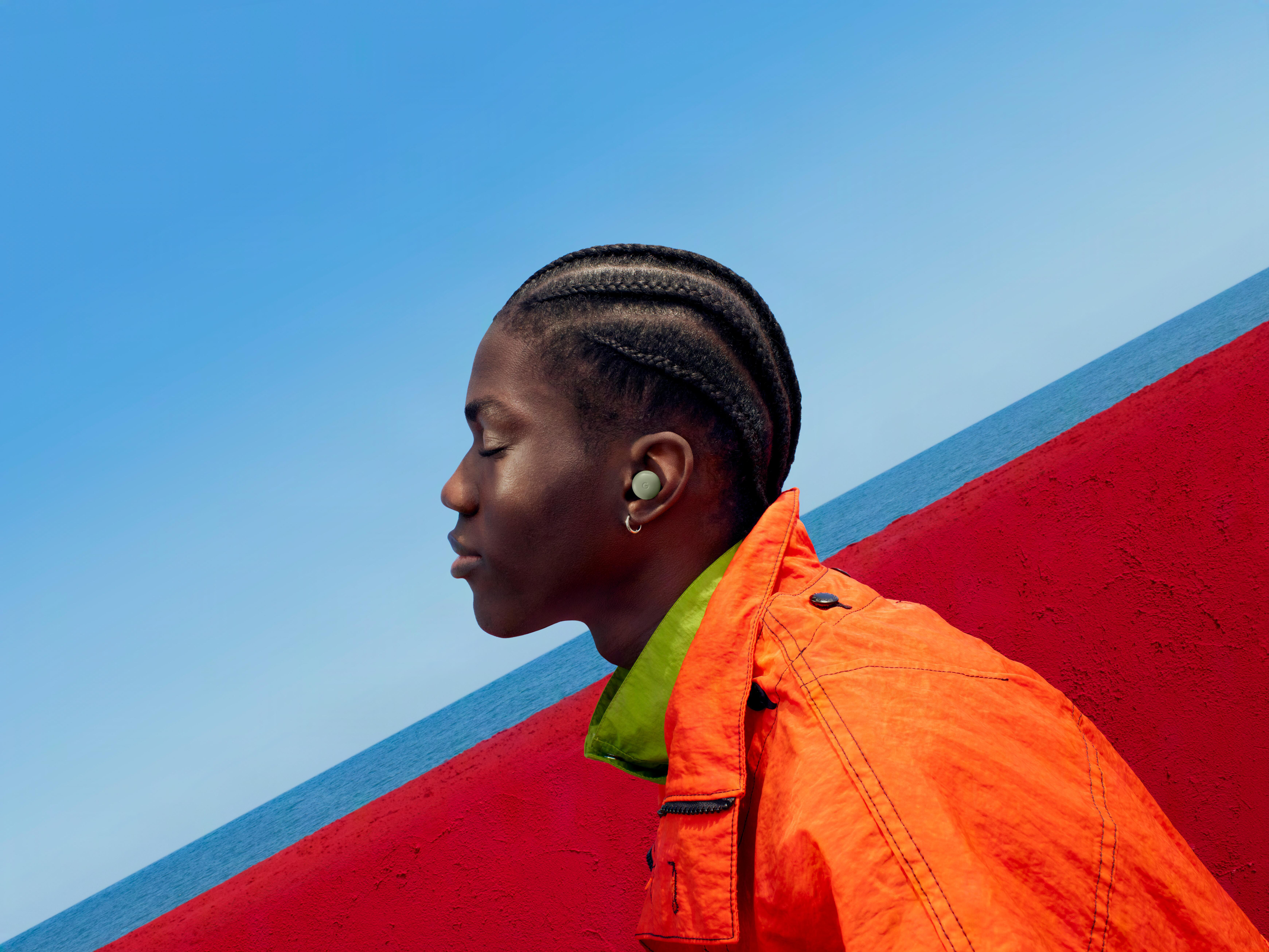 Eine Person mit geschlossenen Augen und Ohrstöpseln, in oranger Jacke vor blauem Himmel und Meer.