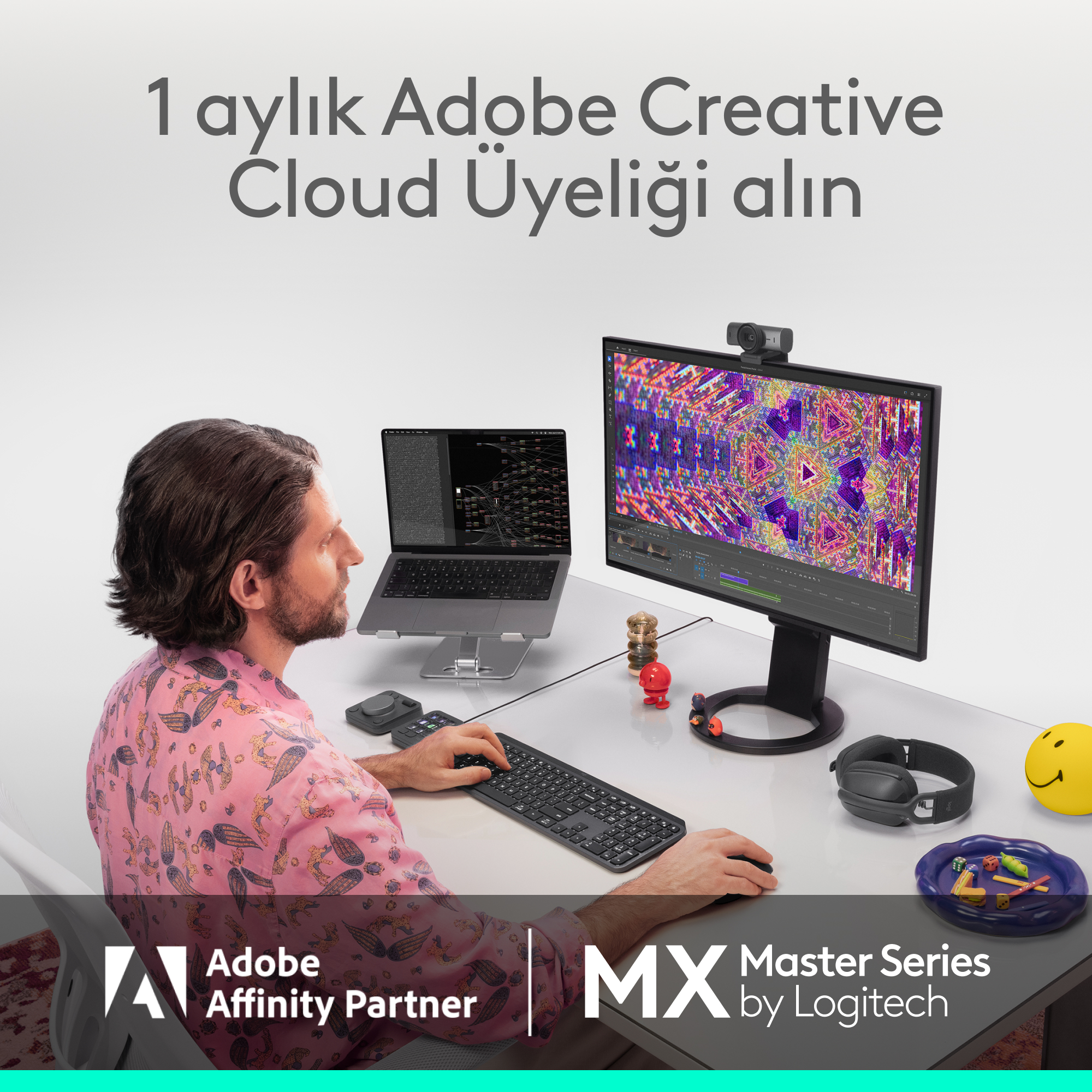 Pembe gömlekli bir adam masada oturarak bilgisayarda çalışıyor; önünde monitör ve dizüstü bilgisayar var.