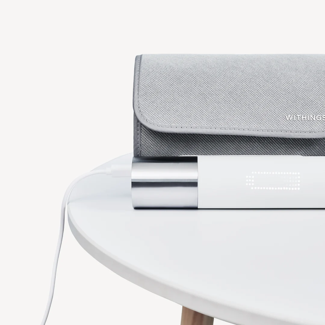 Ein weißes Withings-Gerät lädt mit angeschlossenem Kabel auf einem runden Tisch, ein graues Etui liegt dahinter.