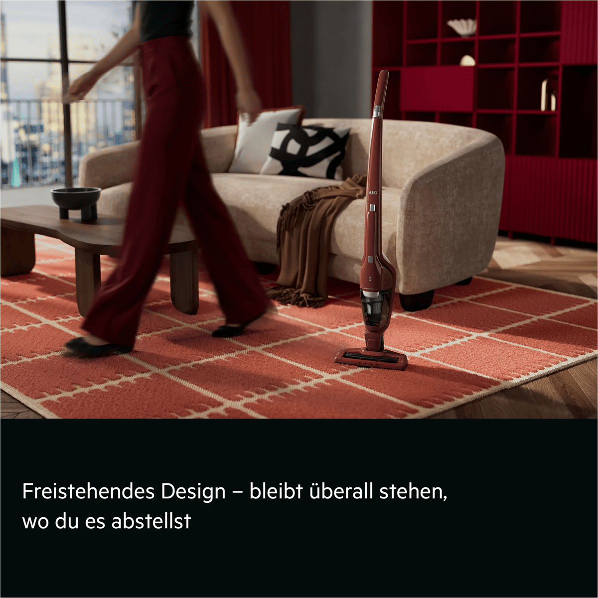 Ein roter Akkustaubsauger steht freistehend auf einem roten Teppich in einem modernen Wohnzimmer.